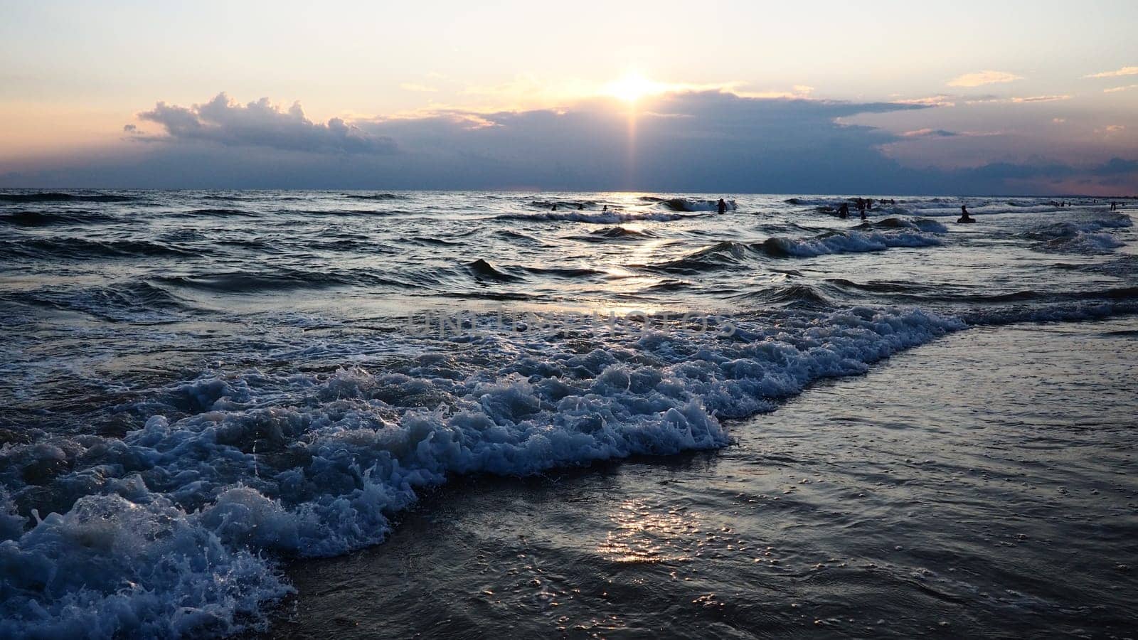 Waves with foam. Beautiful sunset. Foamy waves roll over the sandy shore. Vityazevo, Anapa, Black Sea. Tourist mecca, health resort. Colorful clouds in the evening sky. The rays reflect off the water.