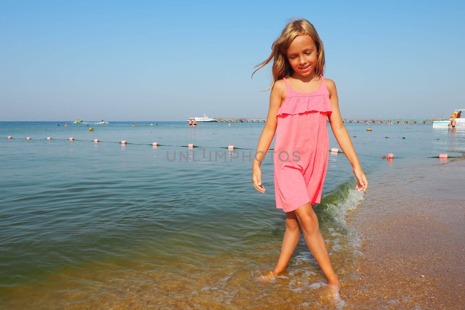 Anapa, Russia, August 29, 2021 A 7-year-old girl in a pink dress walks along the sea. Blond hair and sunburn on the skin. The girl smiles and looks down. Sea wave, swimming people, boats and blue sky.