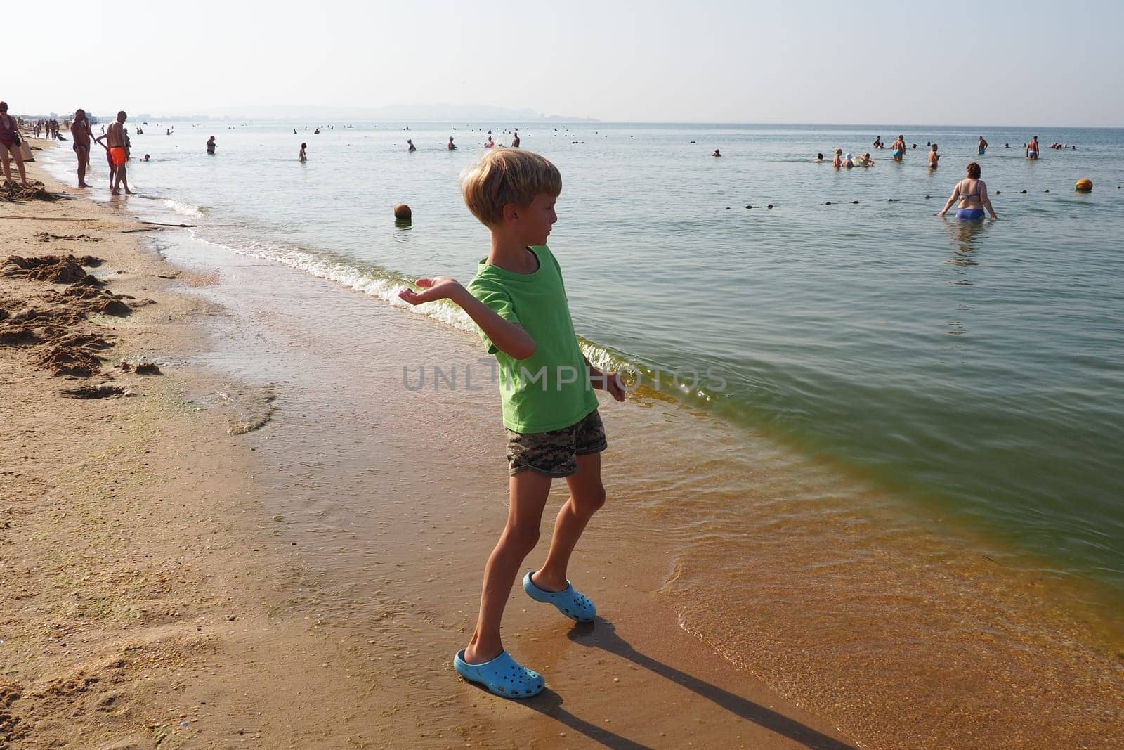Anapa, Russia, August 29, 2021. A 9-year-old boy throws a stone, shell or coin into the sea. Caucasian child with blond hair in green clothes. Sun, sky, sea and swimming tourists. Throwing an object