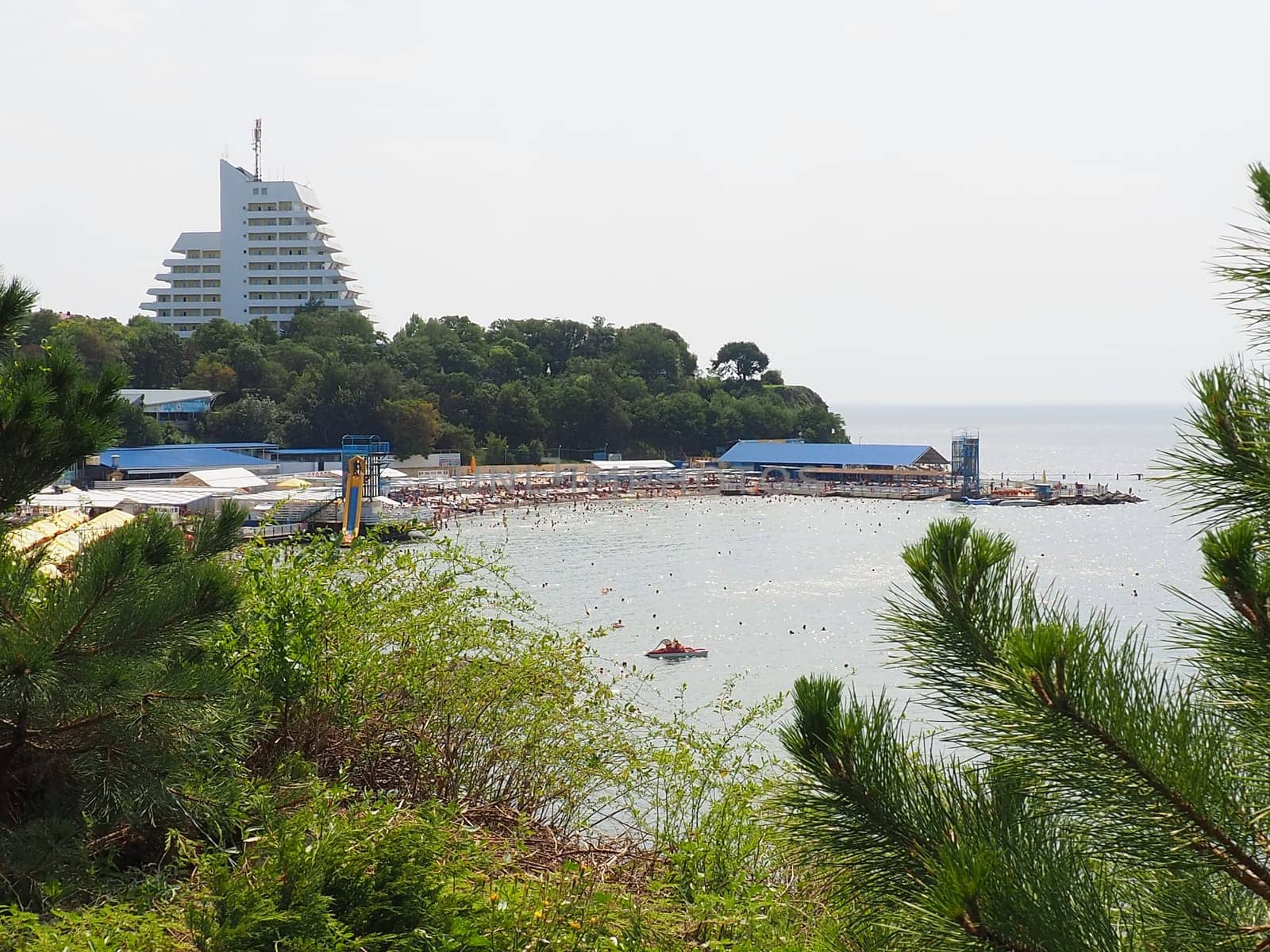 Anapa, Russia, August 23, 2021 Vysokiy Bereg beach or Kamenka. Stone pebbles on the beach. Tourists swim and sunbathe. Summer day. Clear water in Black Sea. Leisure and tourism. Pine branches.