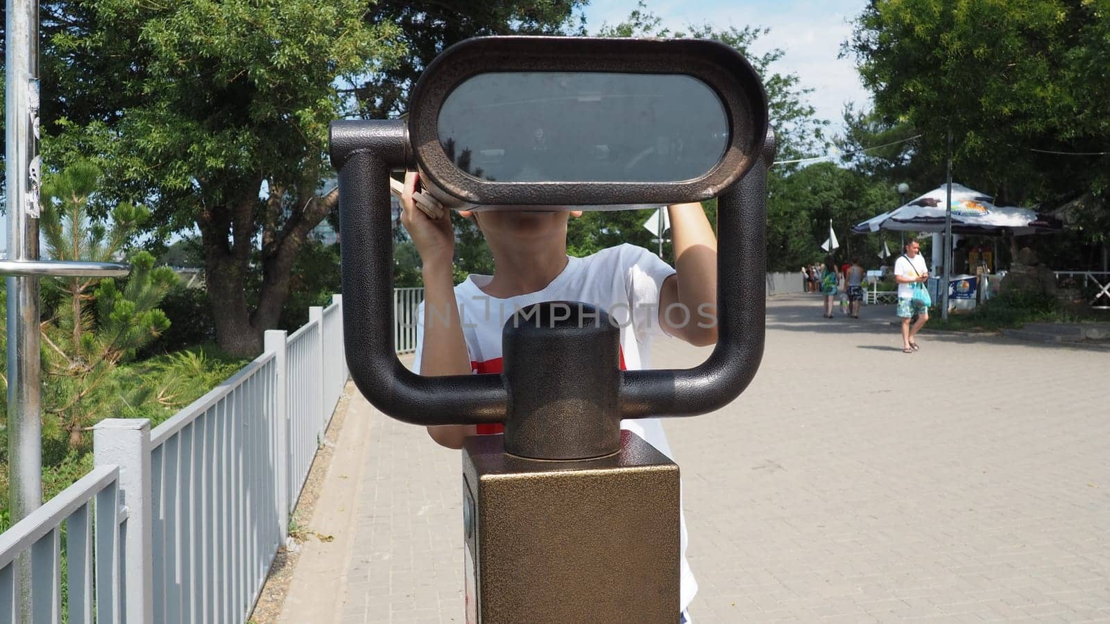 Anapa, Russia, August 23, 2021 A boy looks through an outdoor telescope. A 9-year-old Caucasian child holds a large marine telescope with his hands. The street of the resort town and walking tourists.