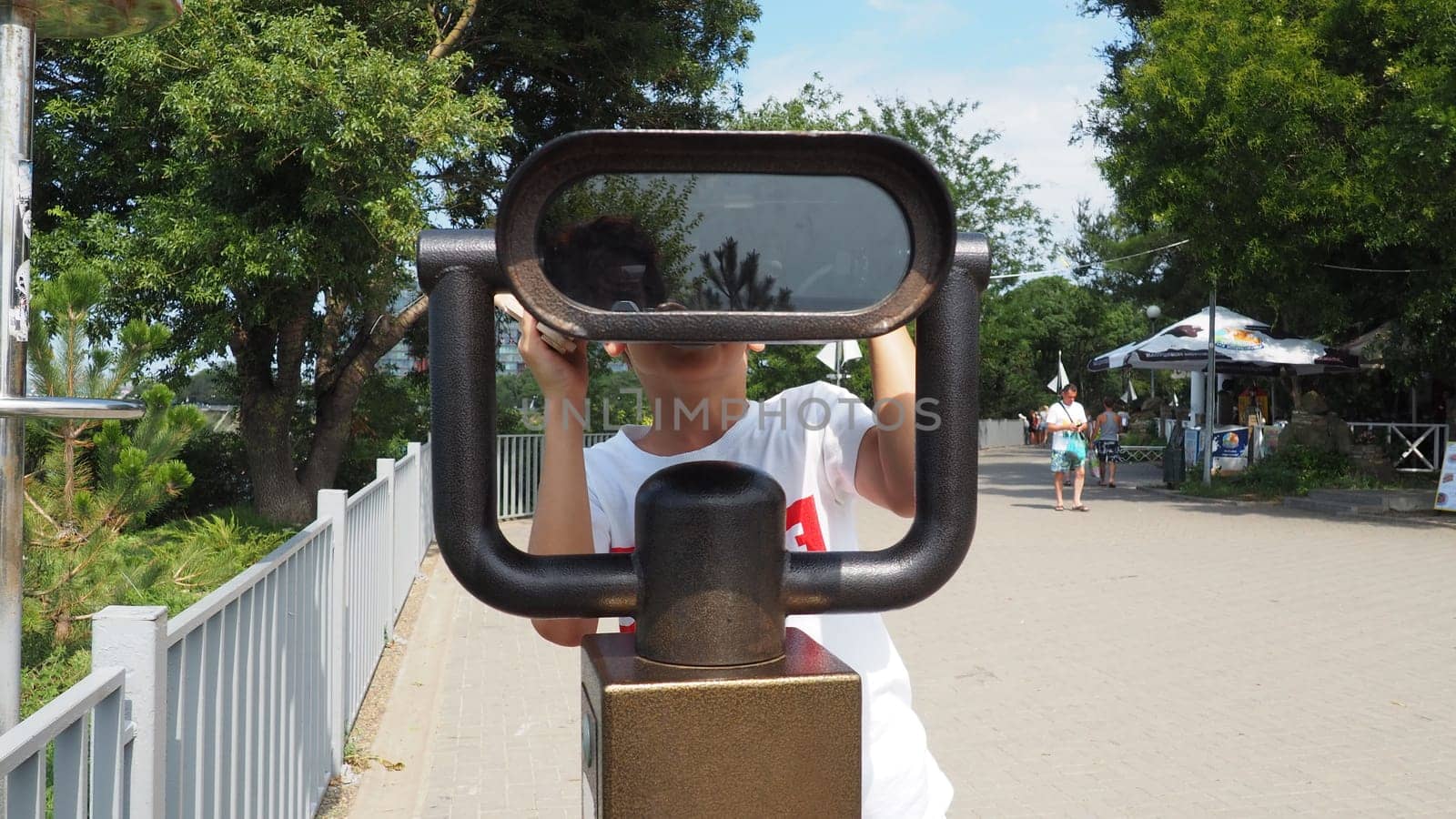 Anapa, Russia, August 23, 2021 A boy looks through an outdoor telescope. A 9-year-old Caucasian child holds a large marine telescope with his hands. The street of the resort town and walking tourists.