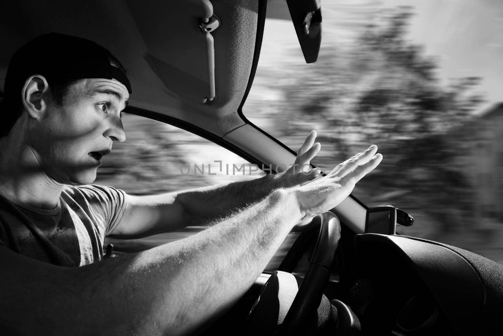 Side wide-angle upper body interior view of a blinded motorist (young man) holding his hands protectively in front of him towards the windshield with shocked look