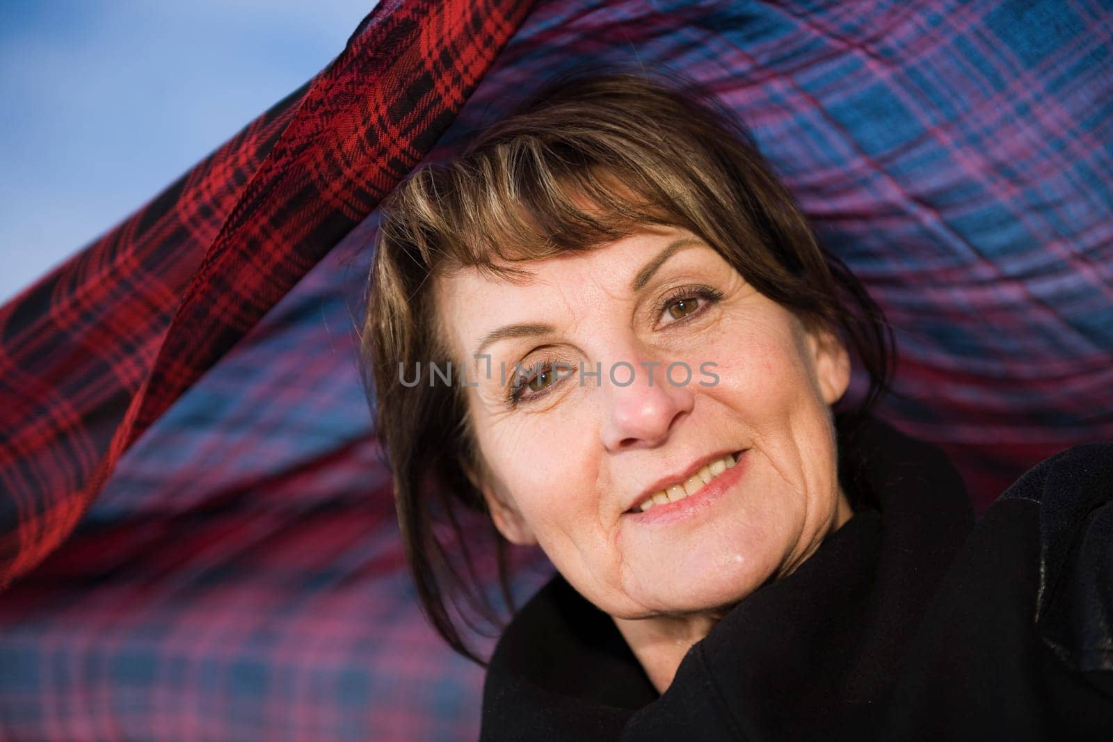 Head portrait of a mature woman in undersight in front of blue sky with a plaid headscarf waving in the wind behind her,smiling friendly at the camera