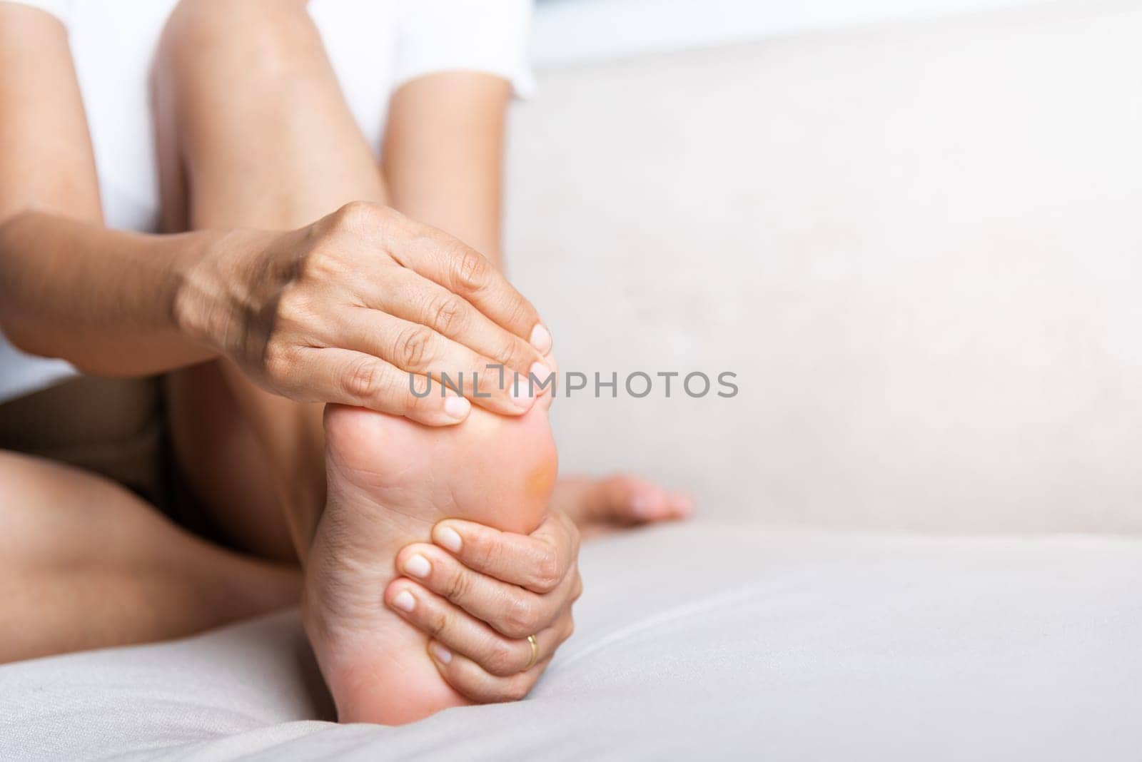Foot pain, Asian woman sitting on sofa feeling pain in her foot at home, female suffering from feet ache use hand massage relax muscle from soles in home interior, Healthcare and podiatry medical