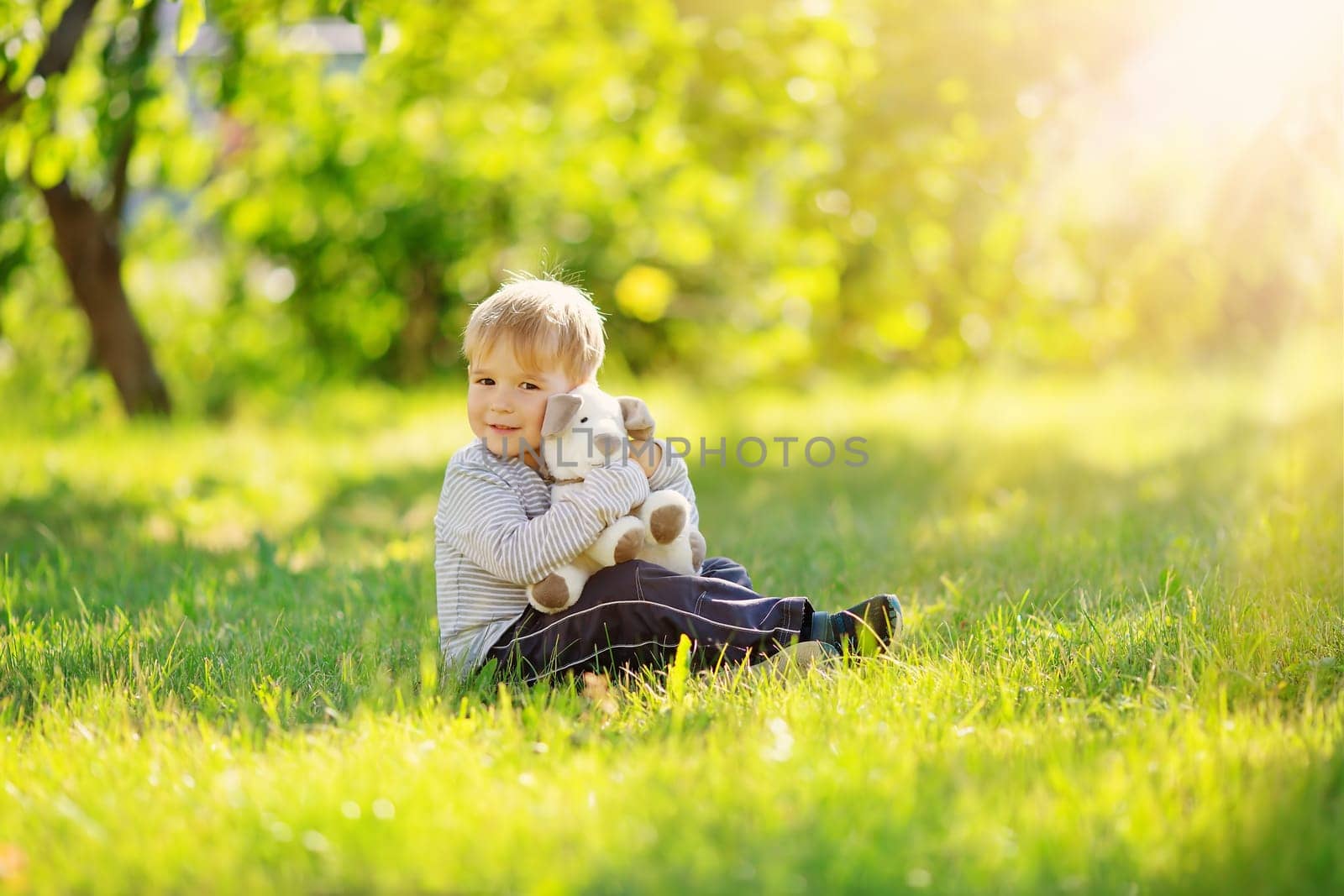 Little cute boy sitting on the meadow in the garden and cuddlying plush toy. Suuny day in nature in spring.