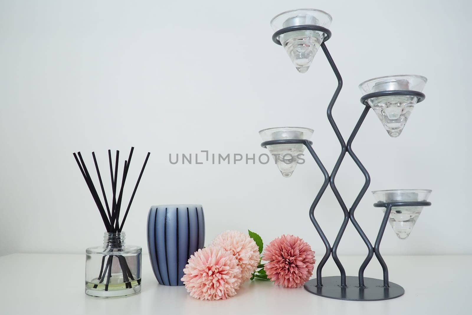 Composition with incense sticks, essential oil in a glass vase and pink terry artificial flowers. Tall metal chandelier with glass holders for small candles. Scented gray striped candle or vase.