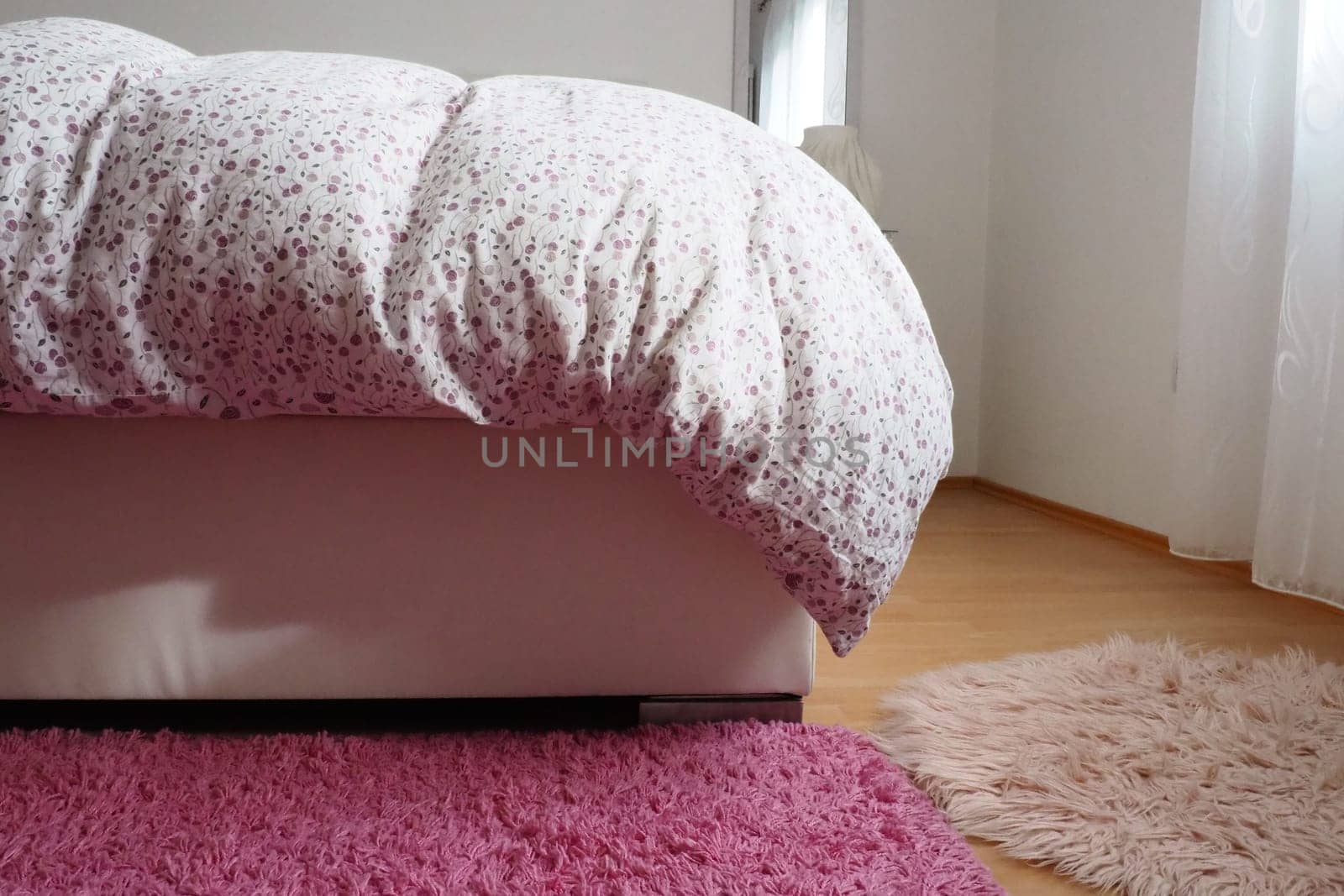 Cozy compact bedroom, white furniture, pink carpet and tulle curtains. Fluffy feather blanket thrown over the double bed. Romantic morning. Light from the window. The blanket hangs down to the floor