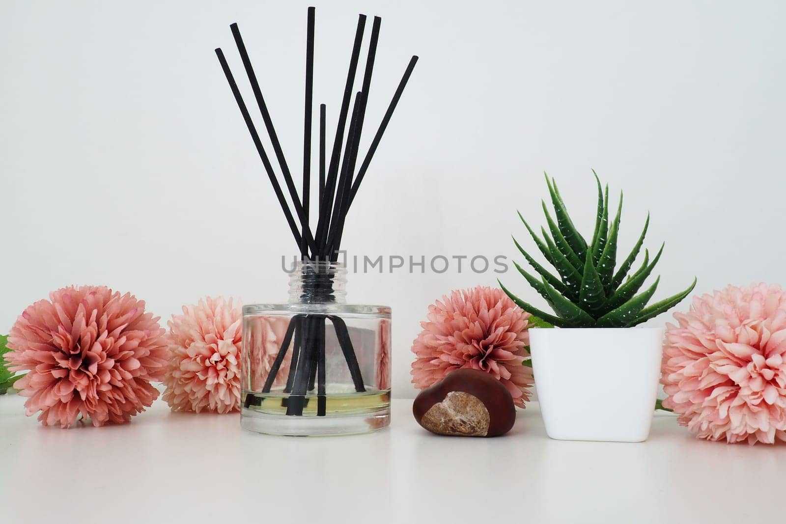 Composition of pink artificial flowers, incense sticks in a glass vase with essential oil. Still life on a white table. Aloe flower in a white pot. Room and apartment decor, interior design