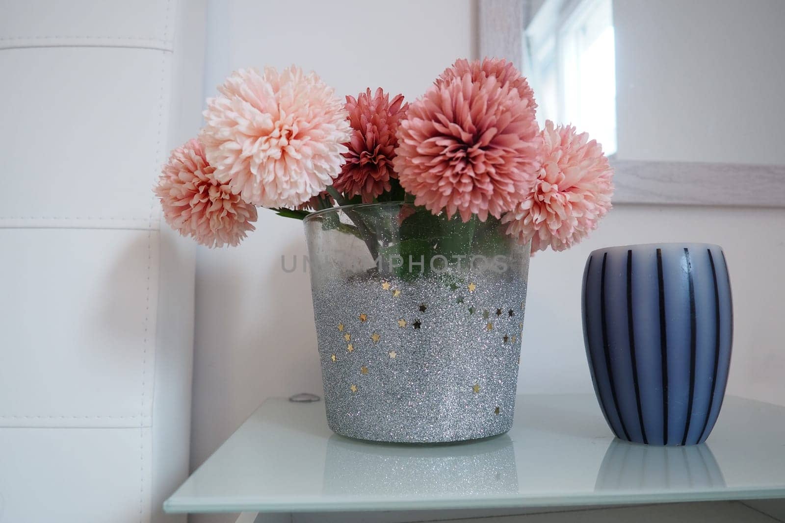 A bouquet of beautiful pink artificial flowers on a white table in the bedroom. Interior Design. Interior design of the women bedroom. Bouquet in a vase. Aromatic striped candle. Mirror on the wall