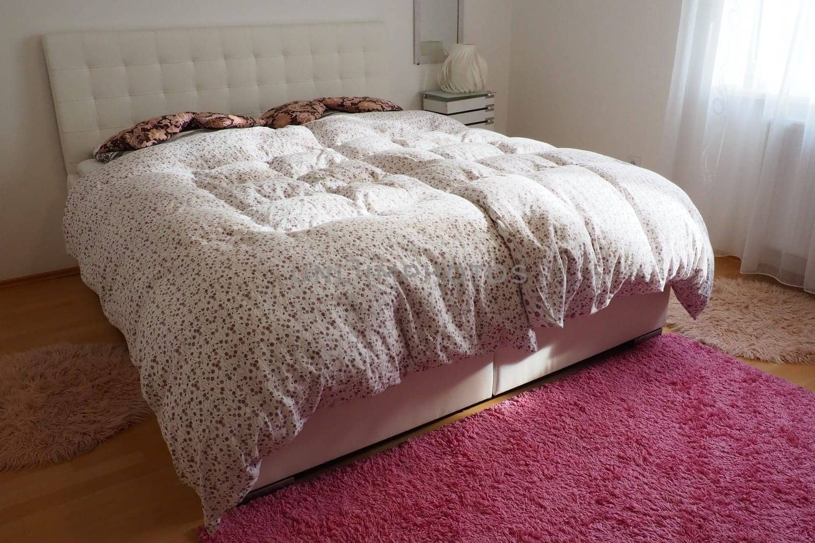 Cozy compact bedroom with white furniture pink carpet and tulle curtains. Fluffy feather blanket is thrown over the bed. Romantic morning. Double bed with upholstered headboard. Light from the window
