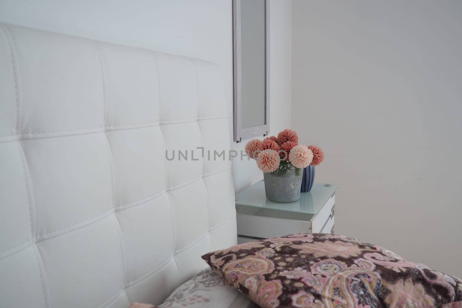 Bedroom interior design. Mirror on the wall. A bouquet of beautiful pink flowers on a white table in the bedroom. Interior Design. Bed with white upholstered headboard. Decorative pillow.