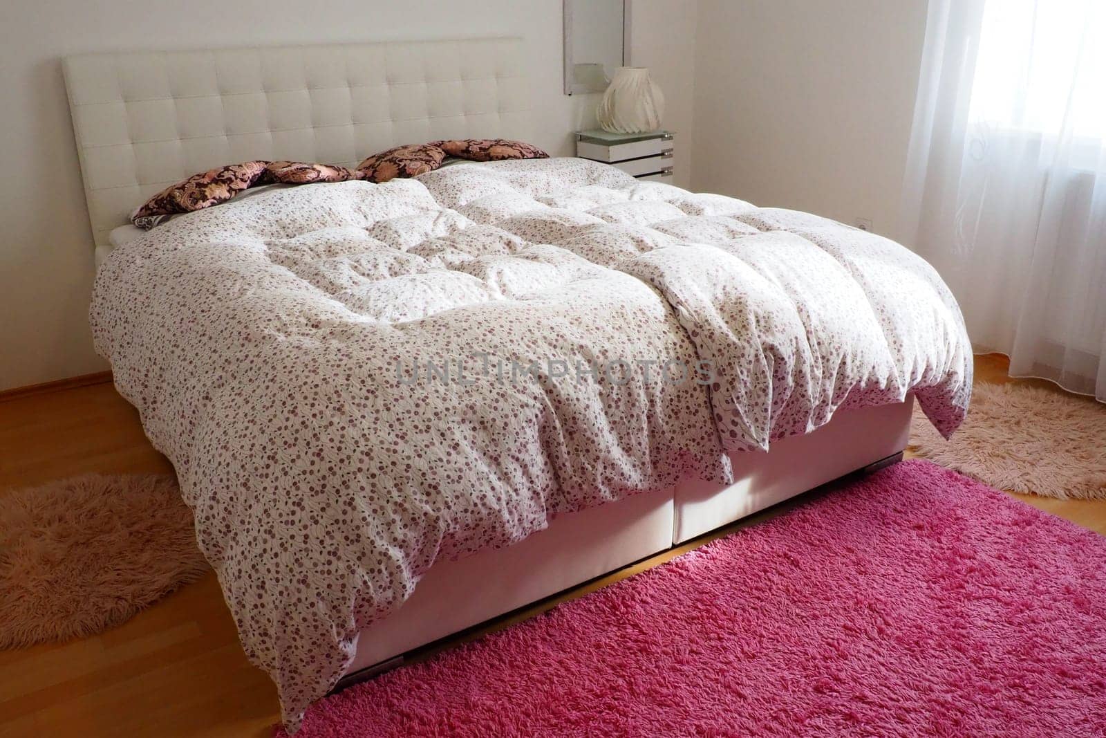 Cozy compact bedroom with white furniture, pink carpet and tulle curtains. Fluffy feather blanket is thrown over the bed. Romantic morning. Double bed with upholstered headboard