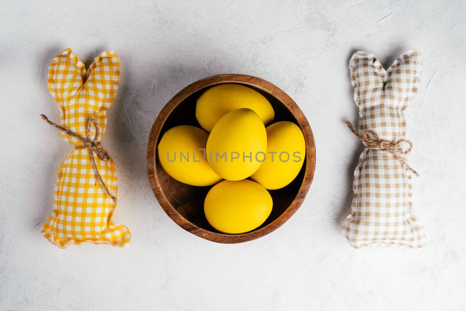 Easter postcard for celebration holiday, two handmade square brown, white and yellow colored bunnies with string-scarf on white gray paper background, yellow chicken eggs in wooden plate