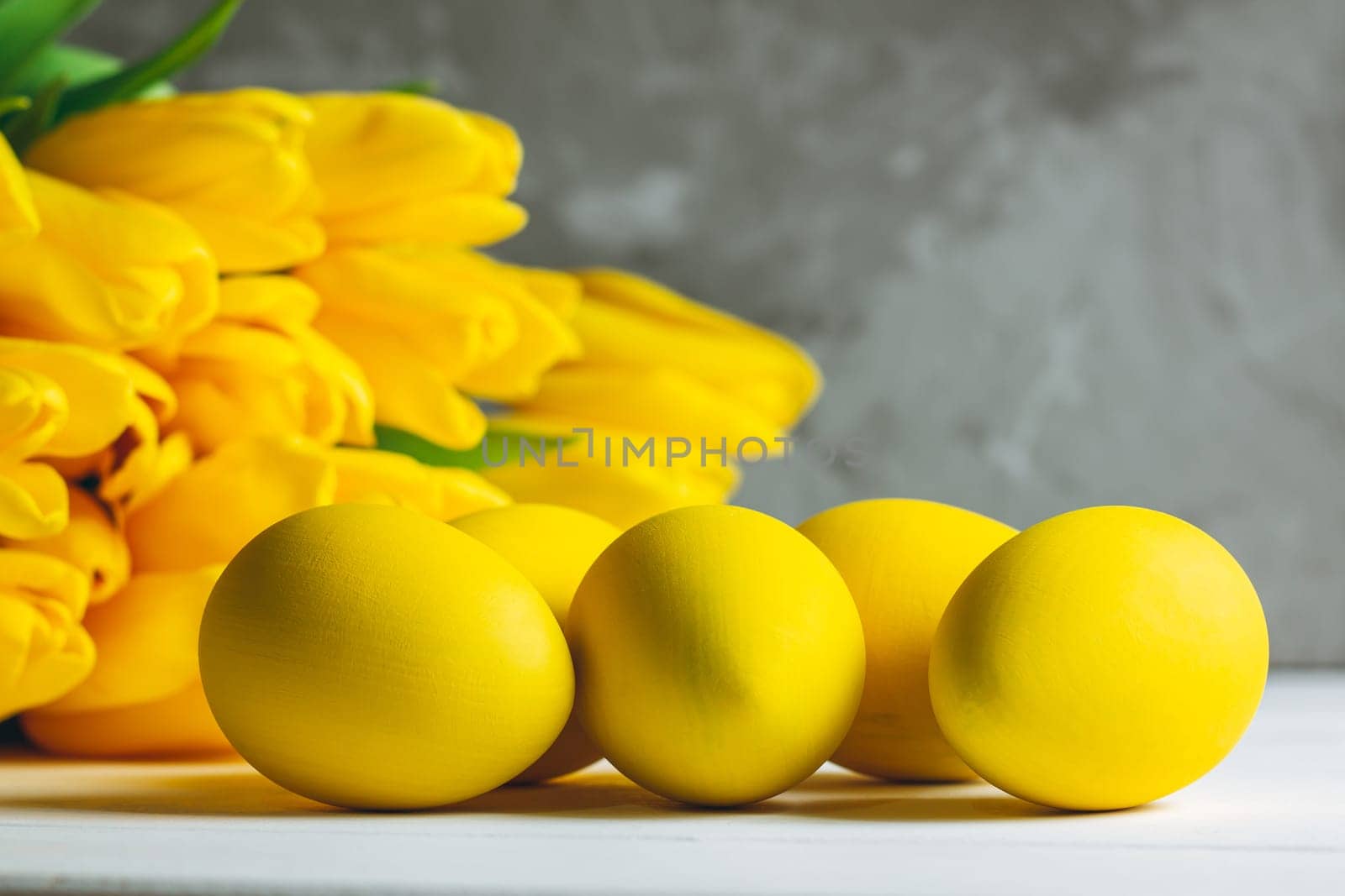 Bouquet of bright yellow tulips and Easter eggs lying on white wooden surface on gray background, copy space
