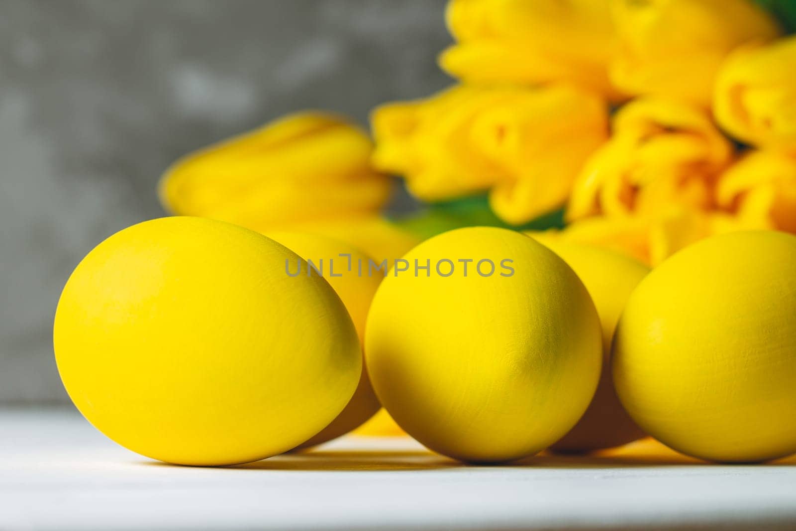 Bouquet of bright yellow tulips and Easter eggs, lying on white wooden surface on gray background