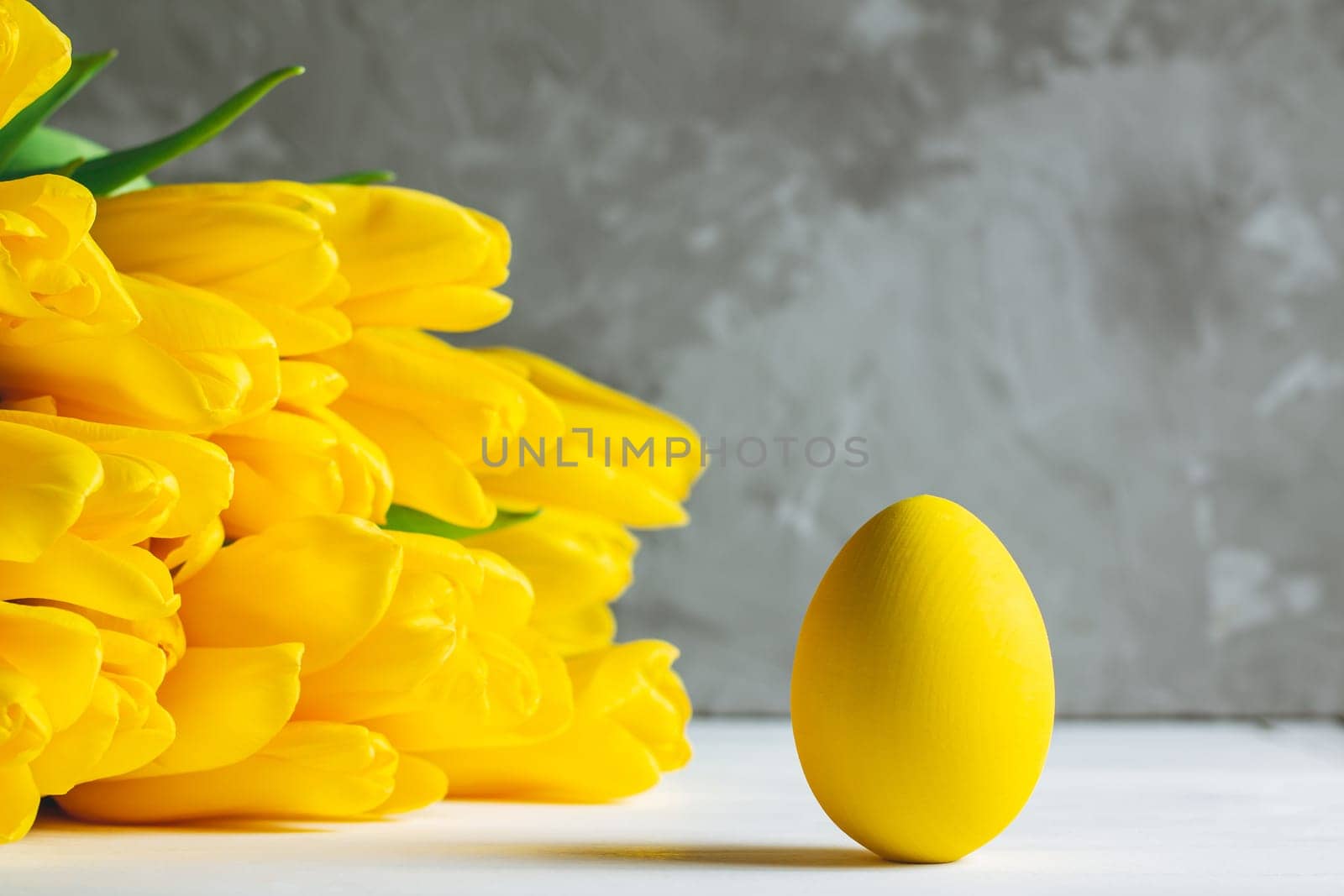 Bouquet of bright yellow tulips and Easter egg, standing on white wooden surface on gray background, copy space