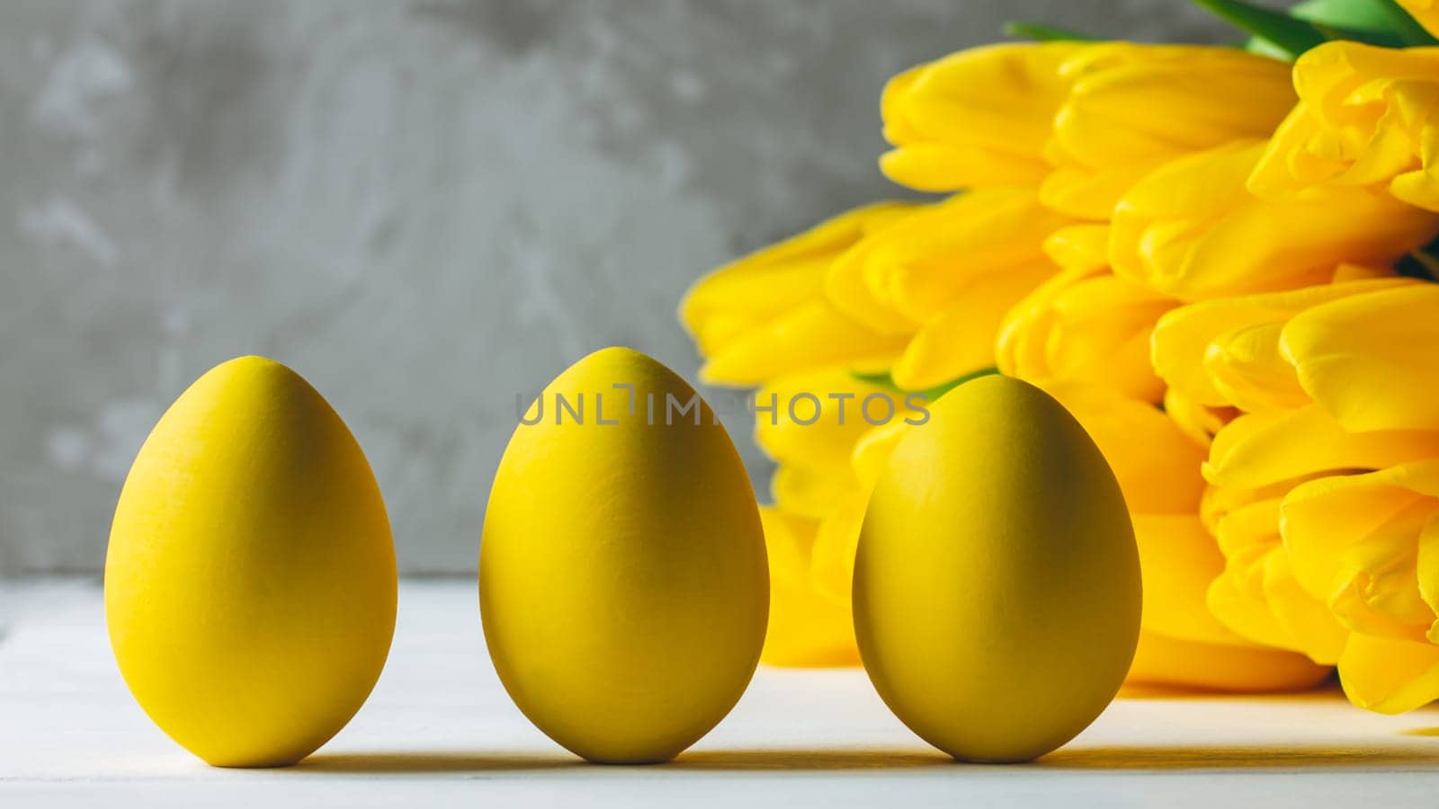 Bouquet of bright yellow tulips and three Easter eggs, standing on white wooden surface on gray background, copy space