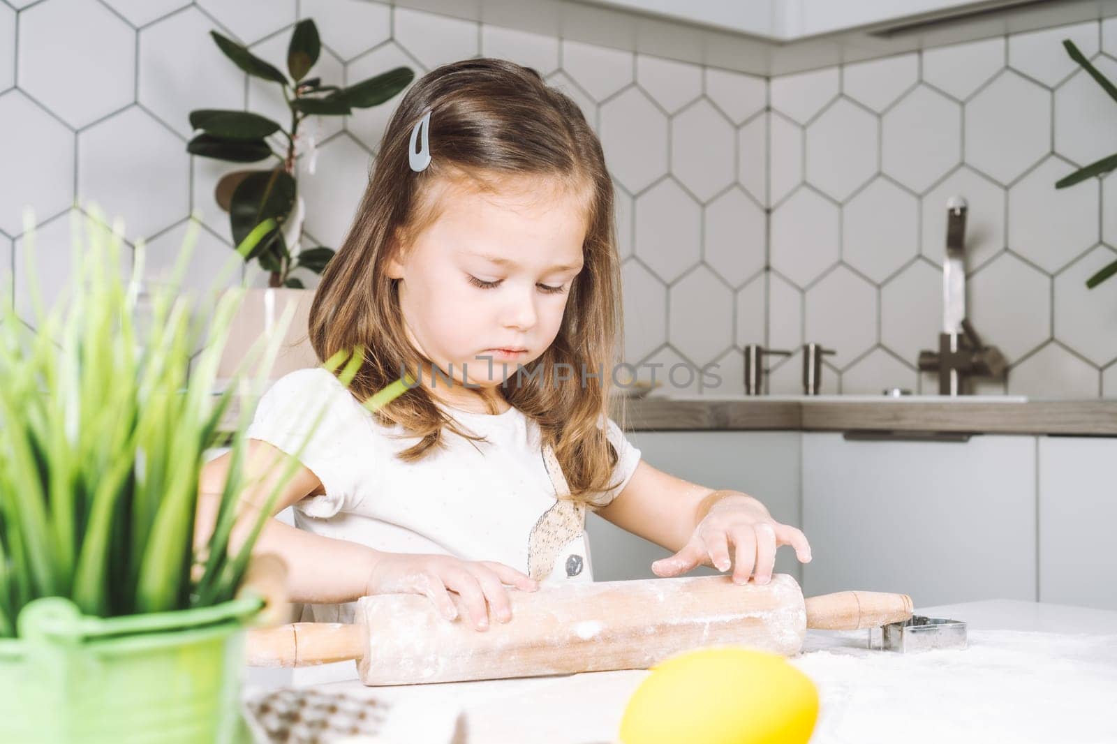 Portrait of little serious girl, helping, learning rolling dough, making shape easter cookies. Sculpt flour biscuit. Preparing holding rolling pin. Yellow egg, plant. Kitchen utensil indoor. Light day