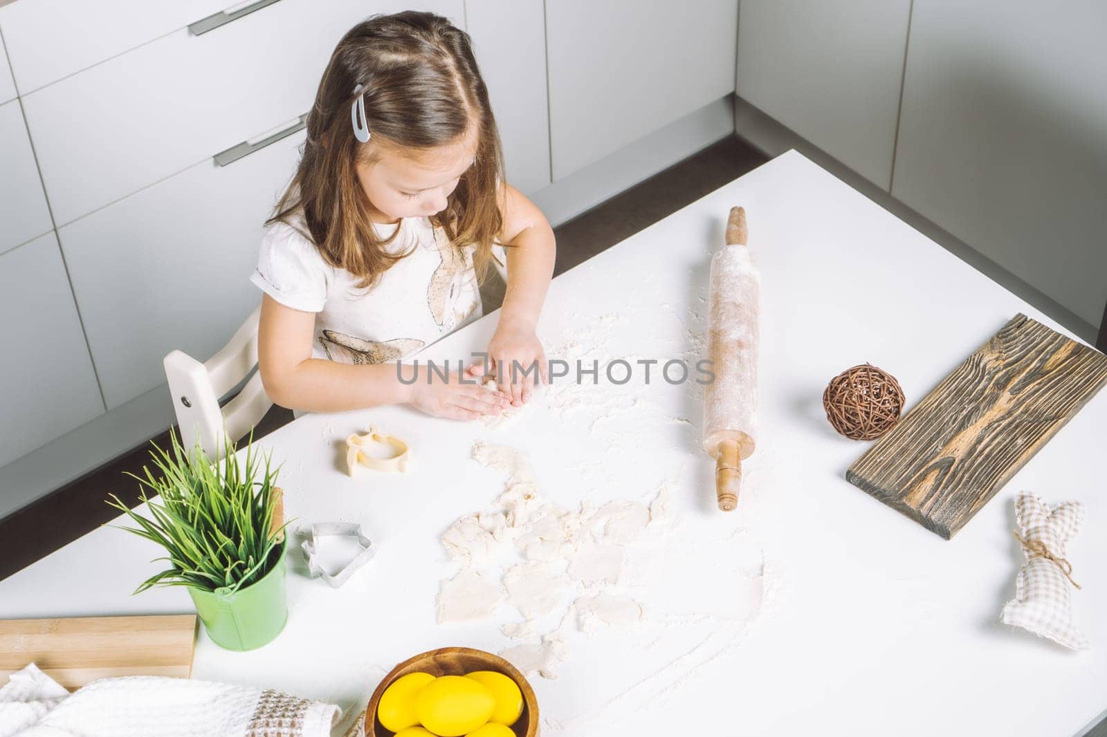 Portrait of little serious girl, helping, making shape dough easter cookies. Sculpt dough flour bakery cup biscuit. Preparing rolling pin. Wood plate of yellow eggs, plant. Kitchen utensil indoor.