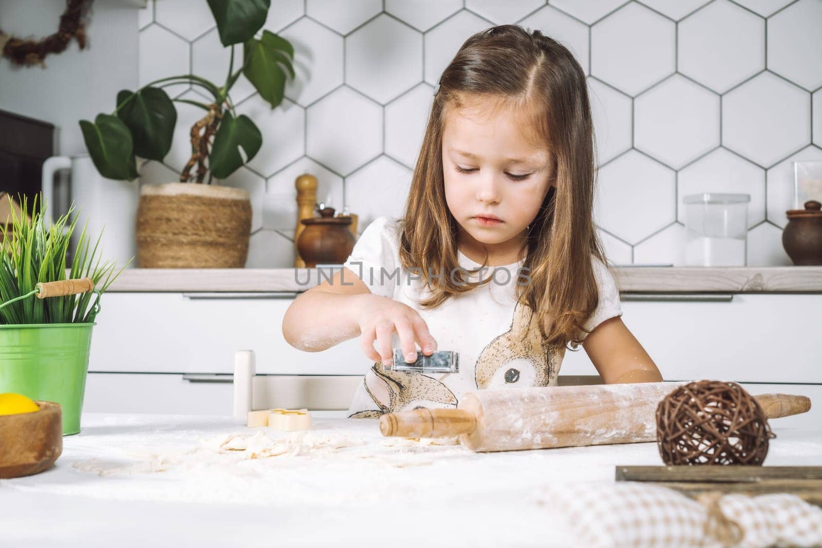 Portrait of little serious girl, helping, making shape dough easter cookies. Sculpt dough flour bakery cup biscuit. Preparing rolling pin. Wood plate of yellow eggs, plant. Kitchen utensil indoor.