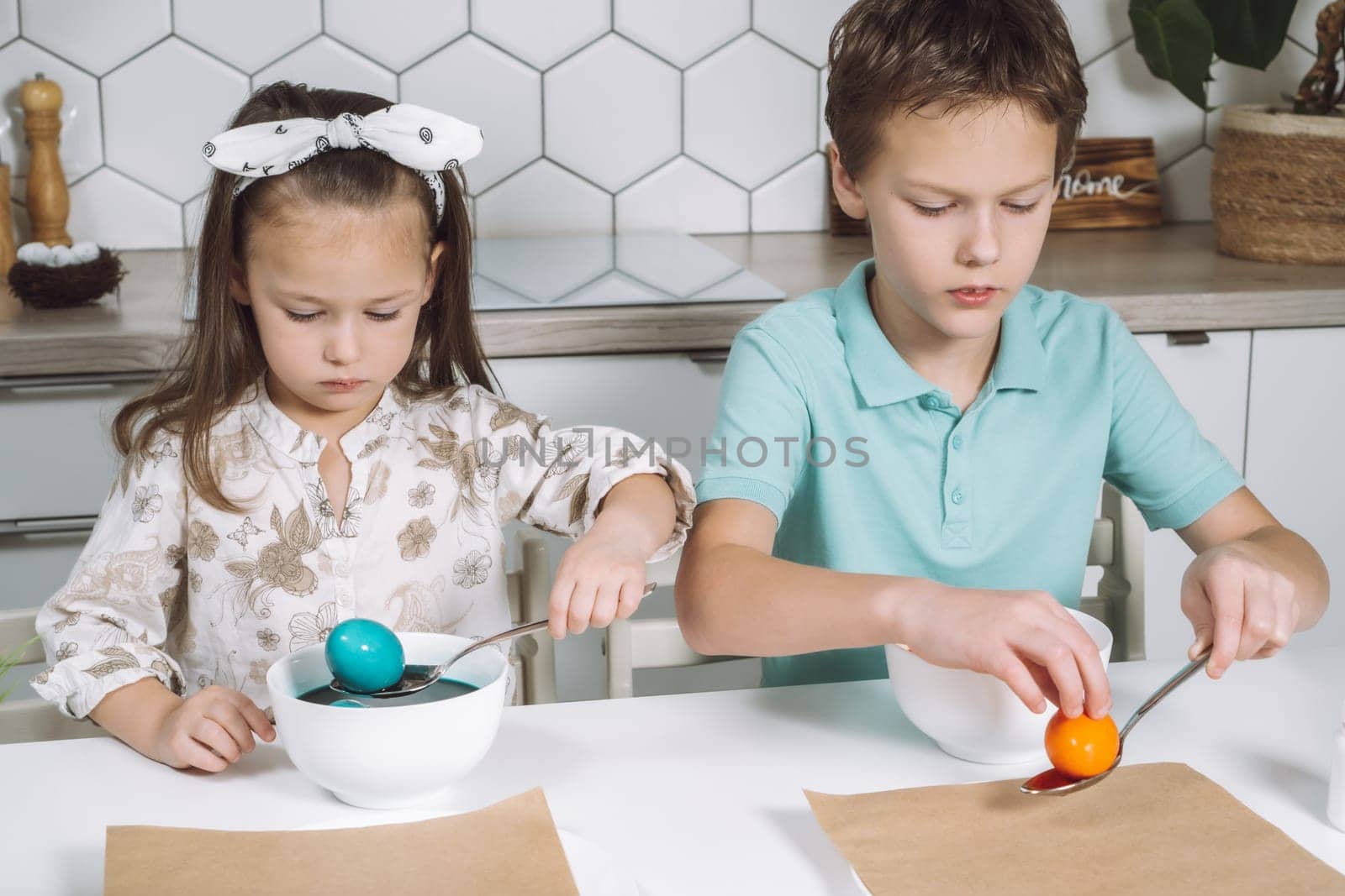 Portrait of concentrated two little children, boy, girl, multi coloring Easter hens eggs. Soaking up, absorption color animal eggs spoon, bowls. Paper on table in kitchen. Traditional holiday event.
