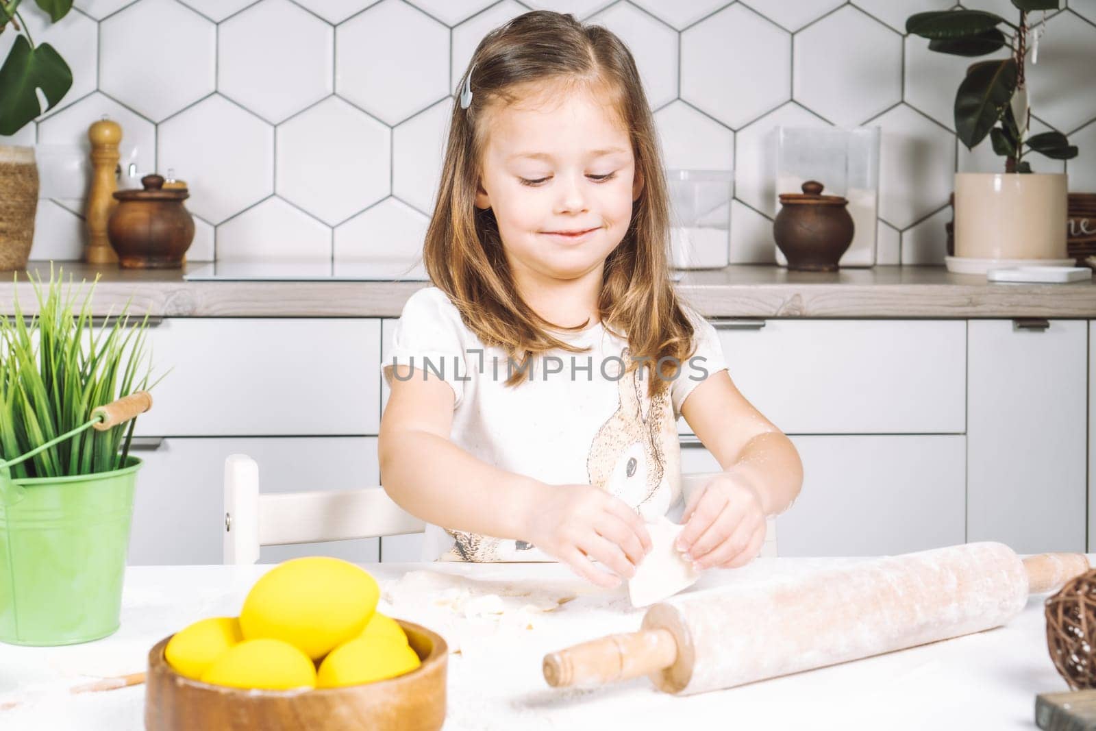 Portrait of little studiously girl sitting on chair, assistance, making different shape cookies. Preparing dough, rolling pin with flour. Sculpting shape biscuit. Plate yellow easter eggs, plant pot