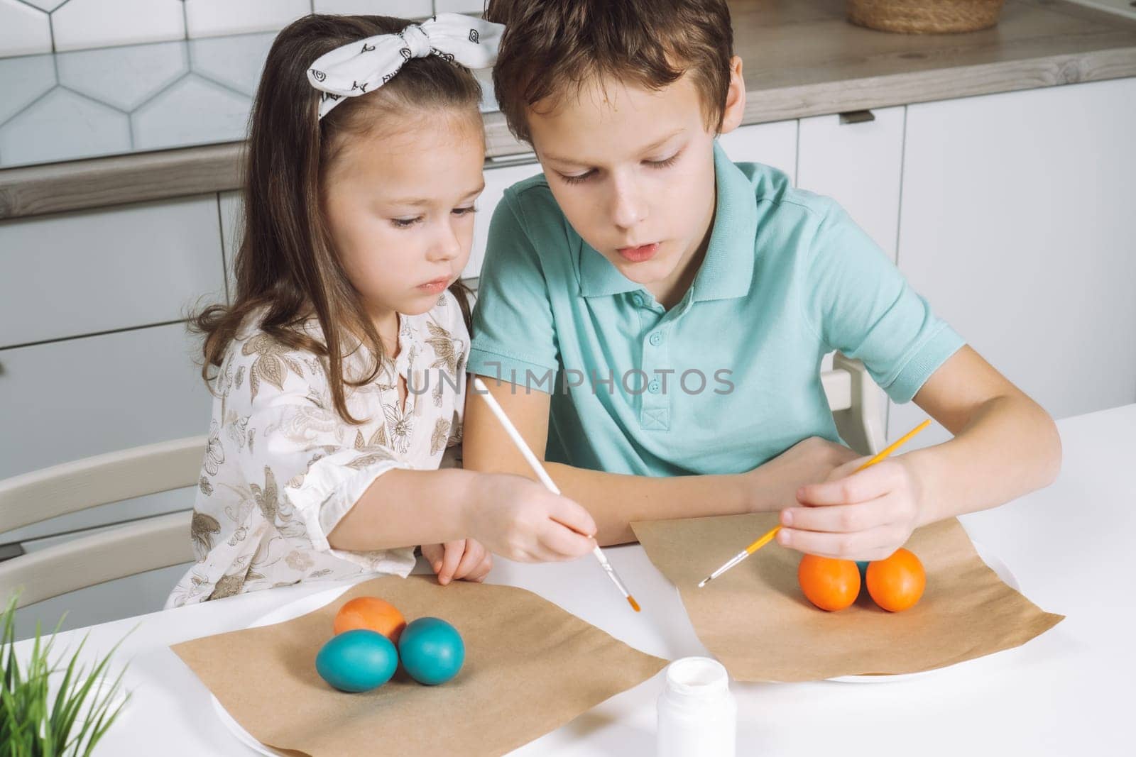 Portrait of enthusiastic two little children, boy and girl, painting brush multi coloring Easter hens eggs paper plate table, plant in kitchen. Making white art patterns. Traditional holiday event.
