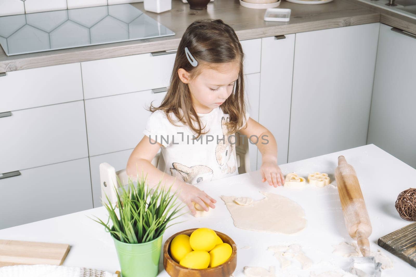 Portrait of concentrated little girl chef, sculpting dough shape cookies in baking cup. Sitting on chair kitchen, Wooden plate of yellow easter eggs and plant pot near child. Kitchen utensil indoor.