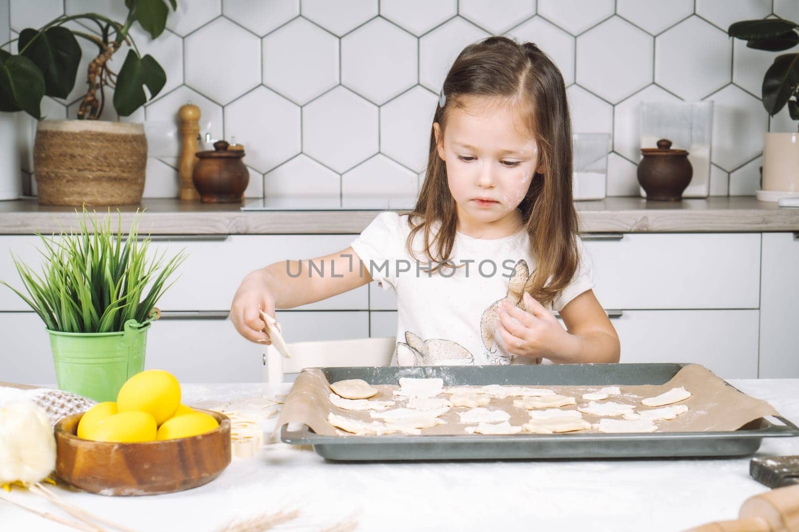 Portrait of little studiously girl chef, sitting on chair, making different shape dough cookies on dripping pan. Wooden plate of yellow easter eggs and plant pot near child. Kitchen utensil indoor.