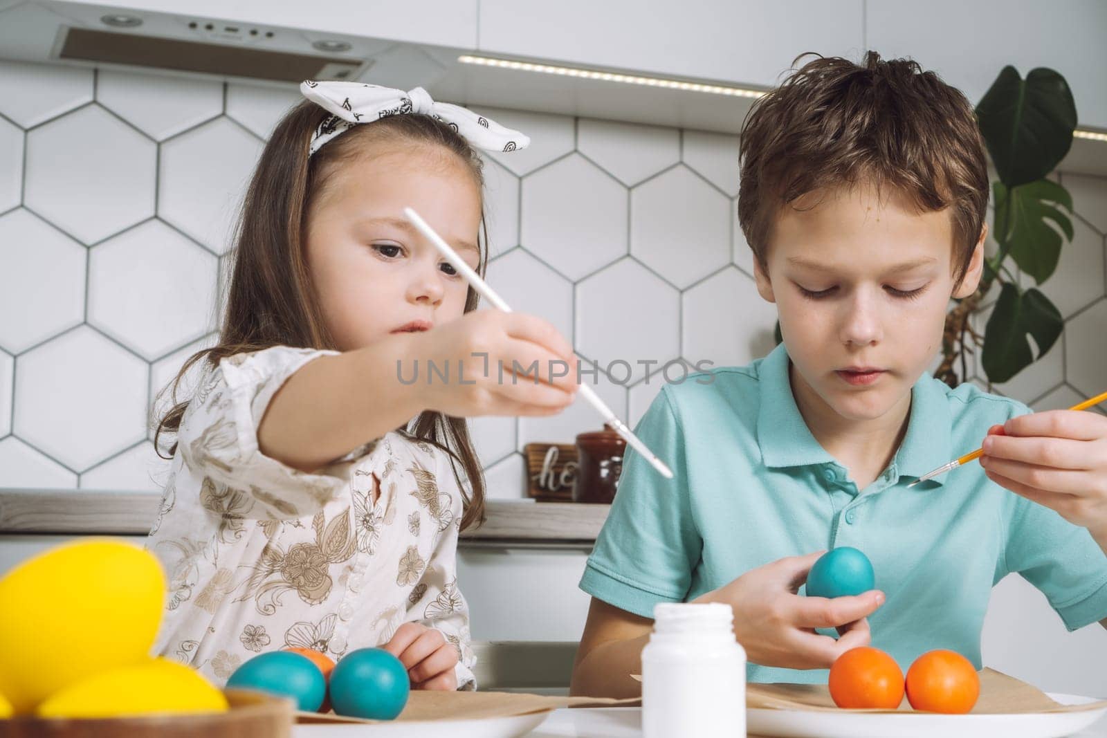 Portrait of enthusiastic two little children, boy and girl, painting brush multi coloring Easter hens eggs paper plate table, plant in kitchen. Making white art patterns. Traditional holiday event.
