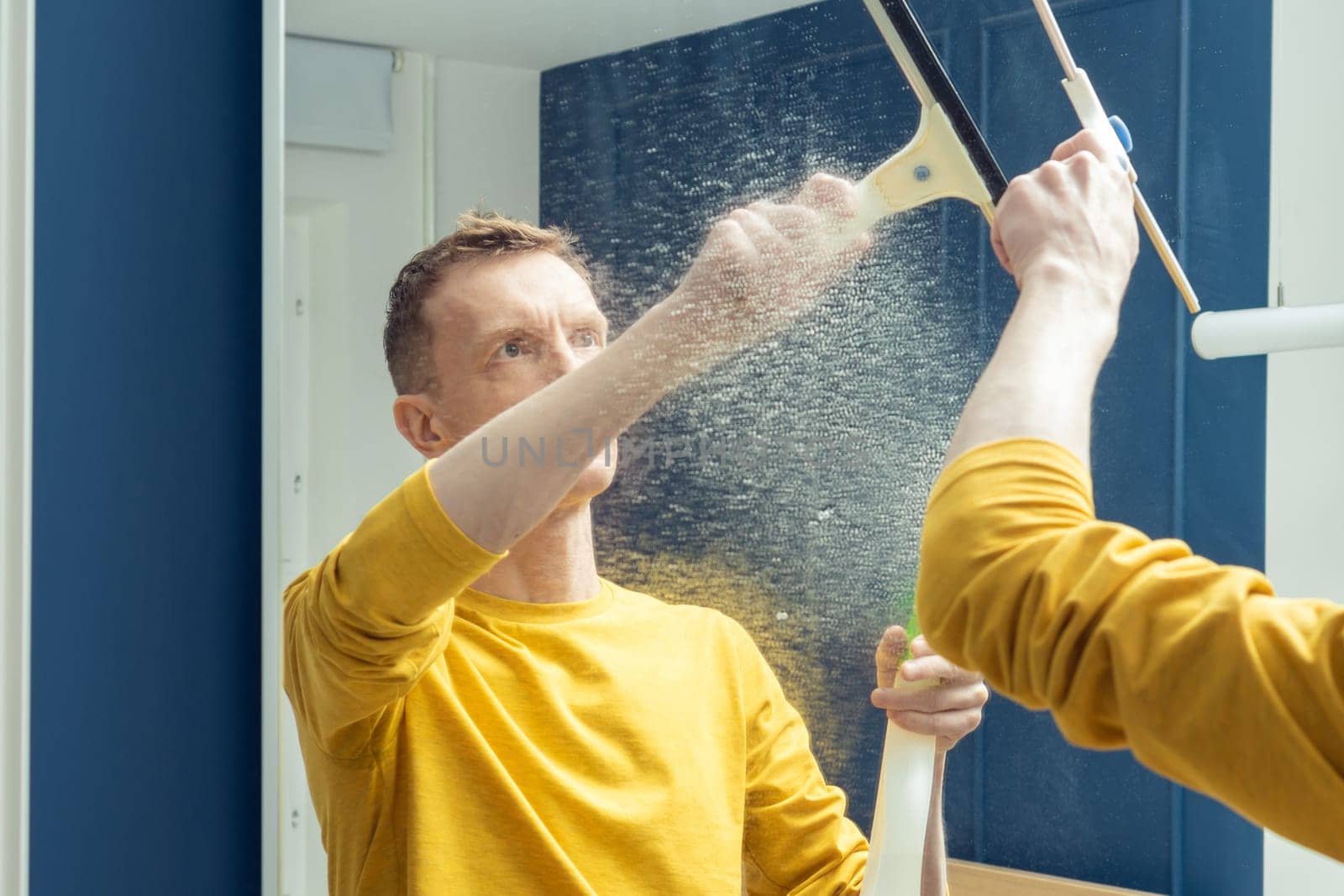 Middle aged man spray cleanser on room mirror and wipe splashes with plastic brush. Rub from dust and polish mirror with household tool. Clear house, housework, wet home cleaning concept.