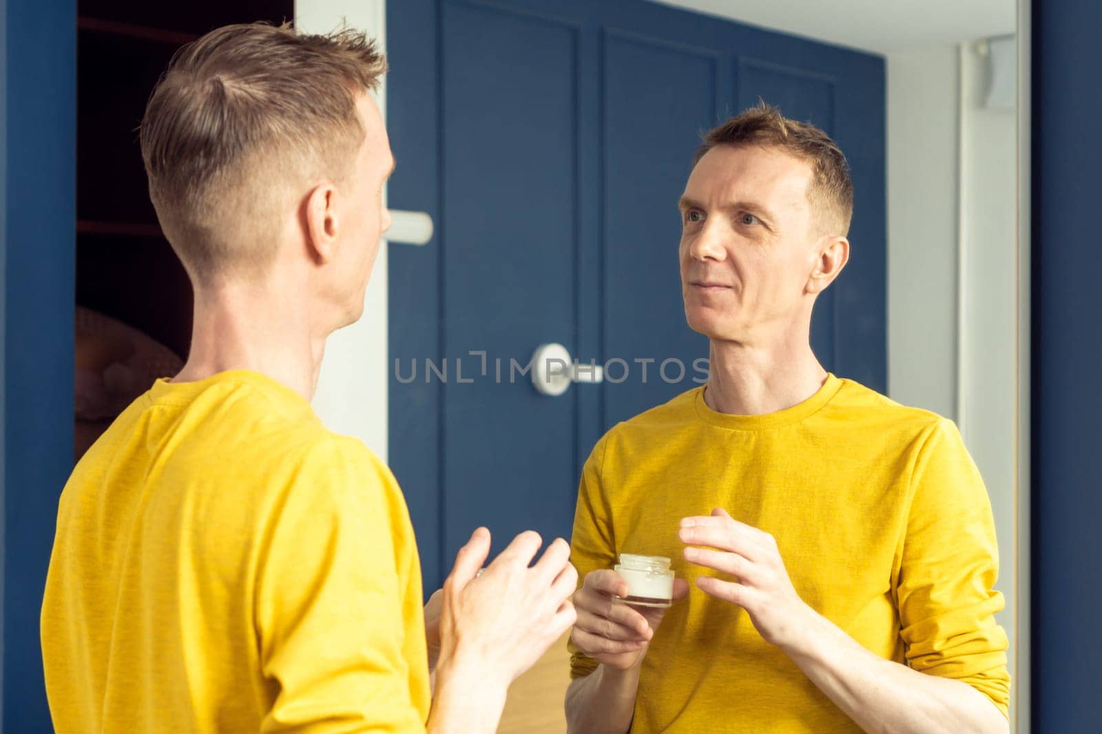 Happy mature man hold jar of moisturizing cream and apply on clear face skin. Male portrait in room mirror. Skincare morning routine, careful beauty treatment, home lifestyle. Facial care concept.