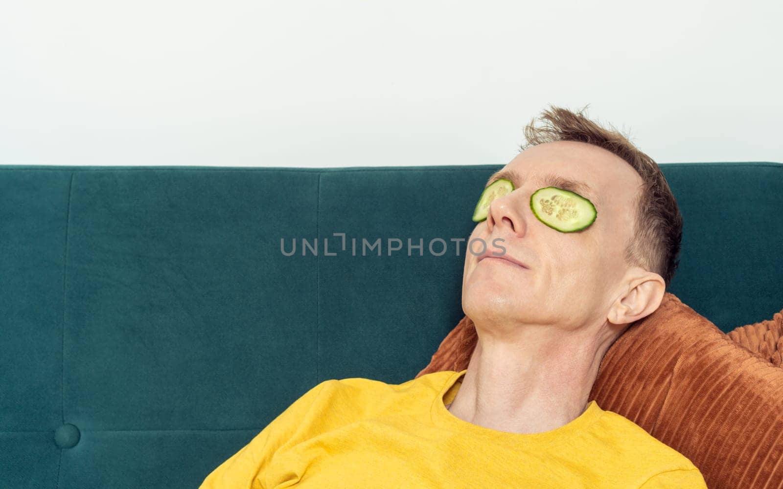 Happy man lying on sofa with eye patches from fresh cucumber slices in living room. Adult person reclining on pillow with mask for dark circles under eyes. Home relaxation, beauty treatment.