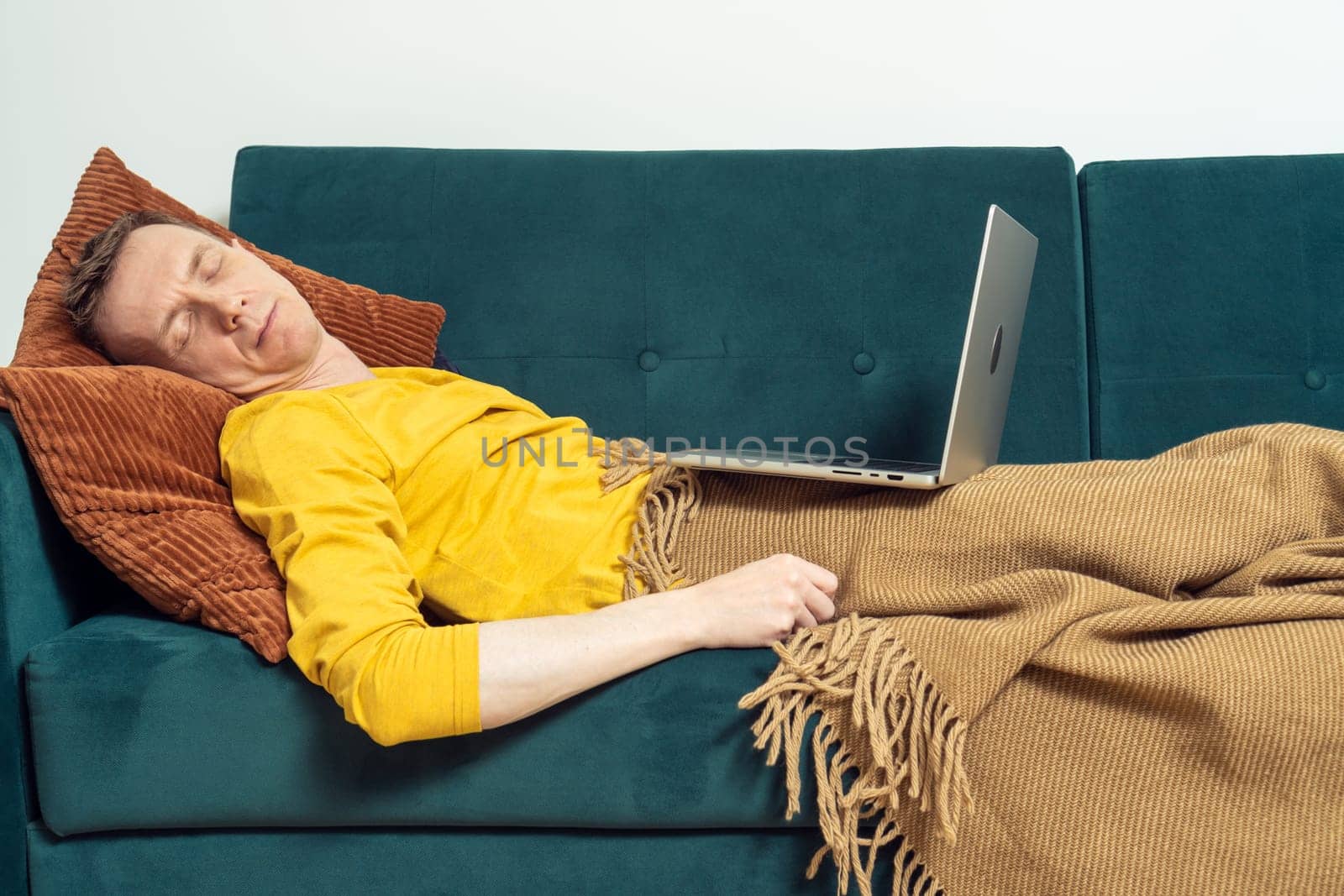 Tired man sleeping on sofa with open laptop in living room. Overworking person reclining on pillow under blanket. Home relaxation, rest break. Business people, remote work, freelancing.