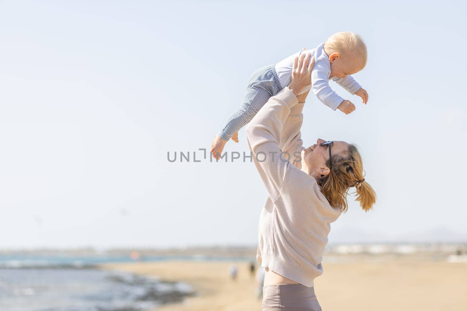 Mother enjoying summer vacations holding, playing and lifting his infant baby boy son high in the air on sandy beach on Lanzarote island, Spain. Family travel and vacations concept