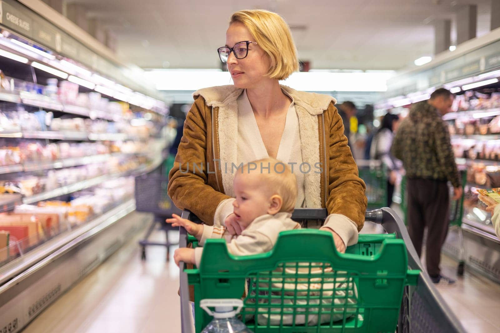 Mother pushing shopping cart with her infant baby boy child down department aisle in supermarket grocery store. Shopping with kids concept