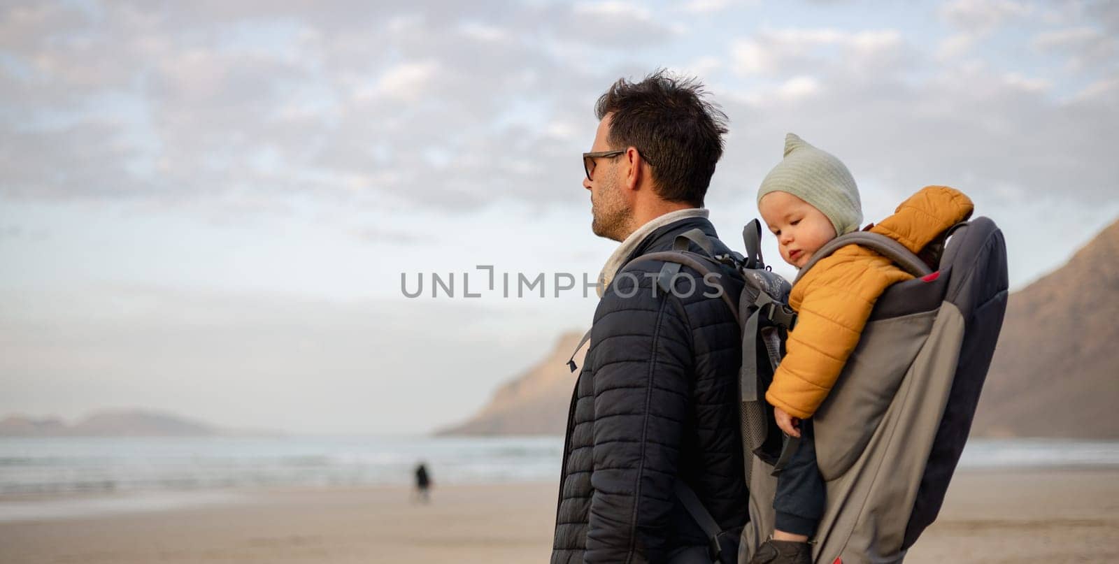 Father rising hands to the sky while enjoying pure nature carrying his infant baby boy son in backpack on windy sandy beach of Famara, Lanzarote island, Spain at sunset. Family travel concept