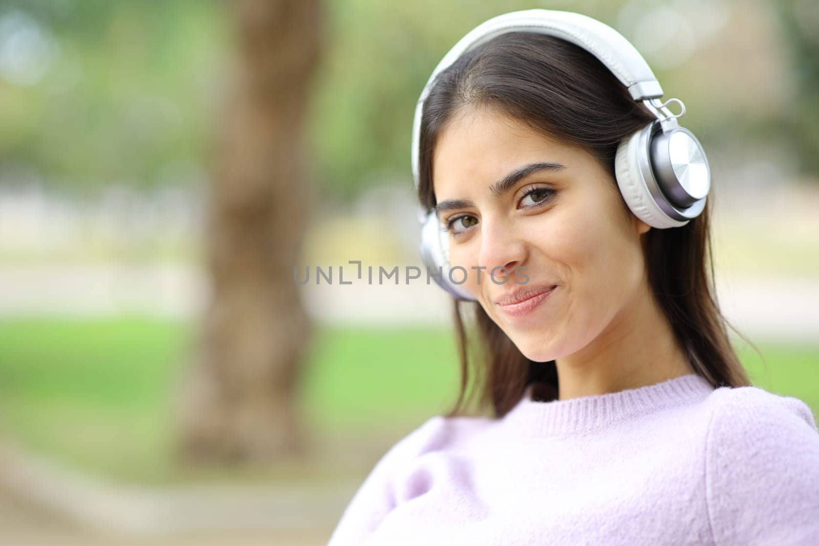 Happy woman with headphones looks at you in a park