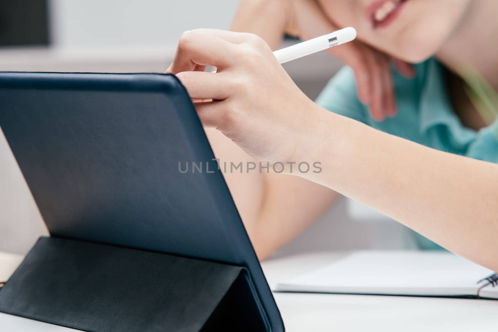 Cropped photo of young preteen boy wearing blue T-shirt, sitting at desk with digital tablet, writing drawing on screen with white pen stylus. Remote education, online school, e-learning, technology.