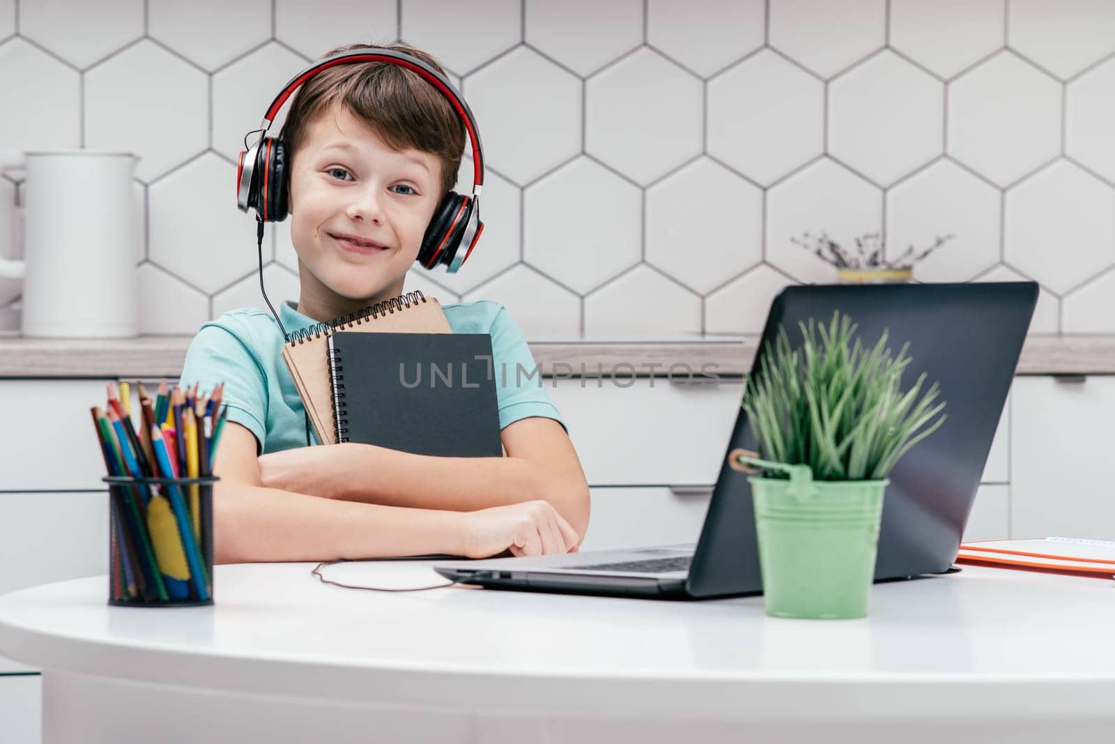 Portrait of young preteen boy wearing blue T-shirt, black red headphones, sitting at desk in front of laptop near houseplant, holding cuddling notebooks. Remote education, technology, e-learning.