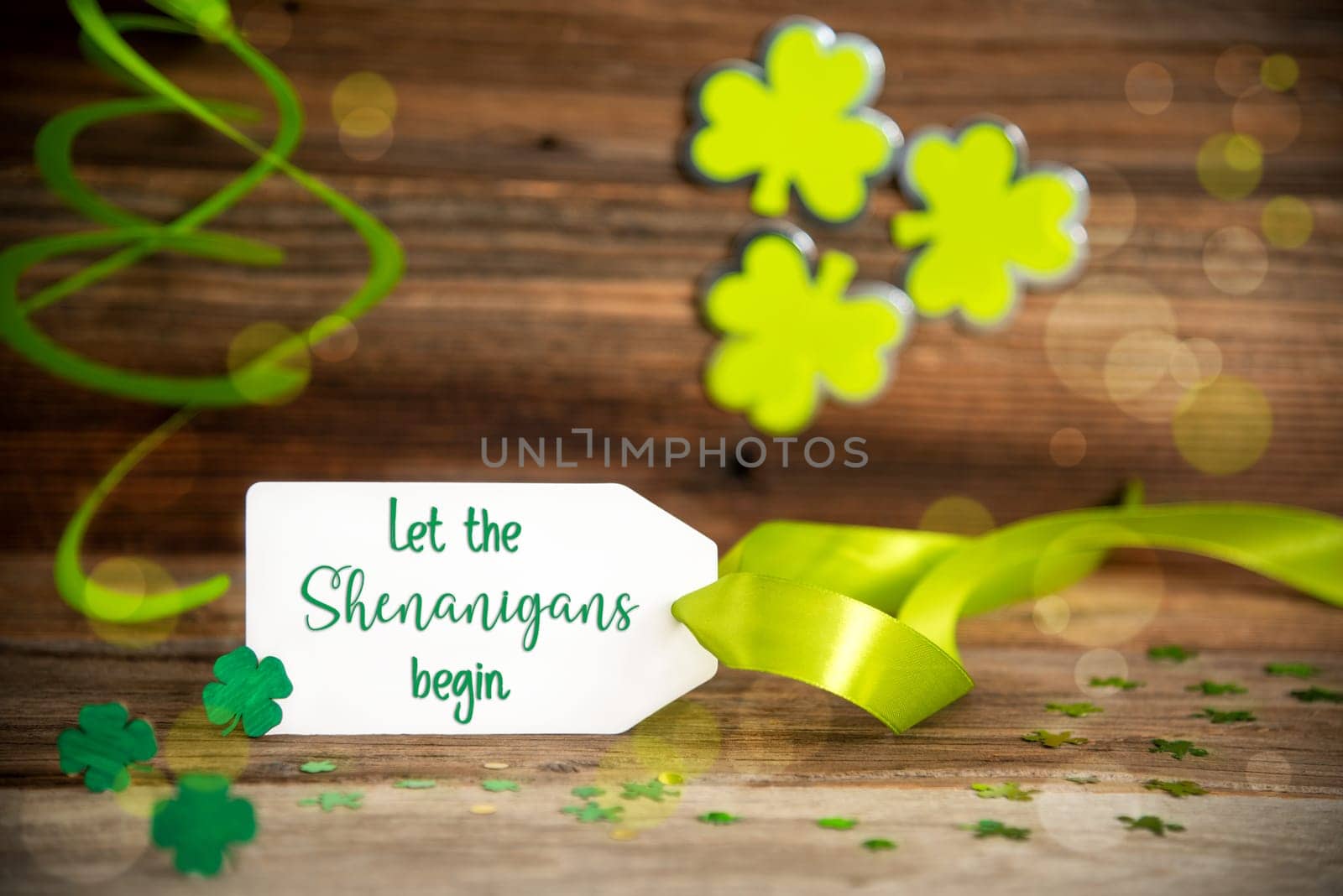 White Label With English Text Let The Shenanigans Begin. Saint Patrick's Day Decoration With Green Clover And Shamrock. Wooden Vintage Background With Festive Ribbon.