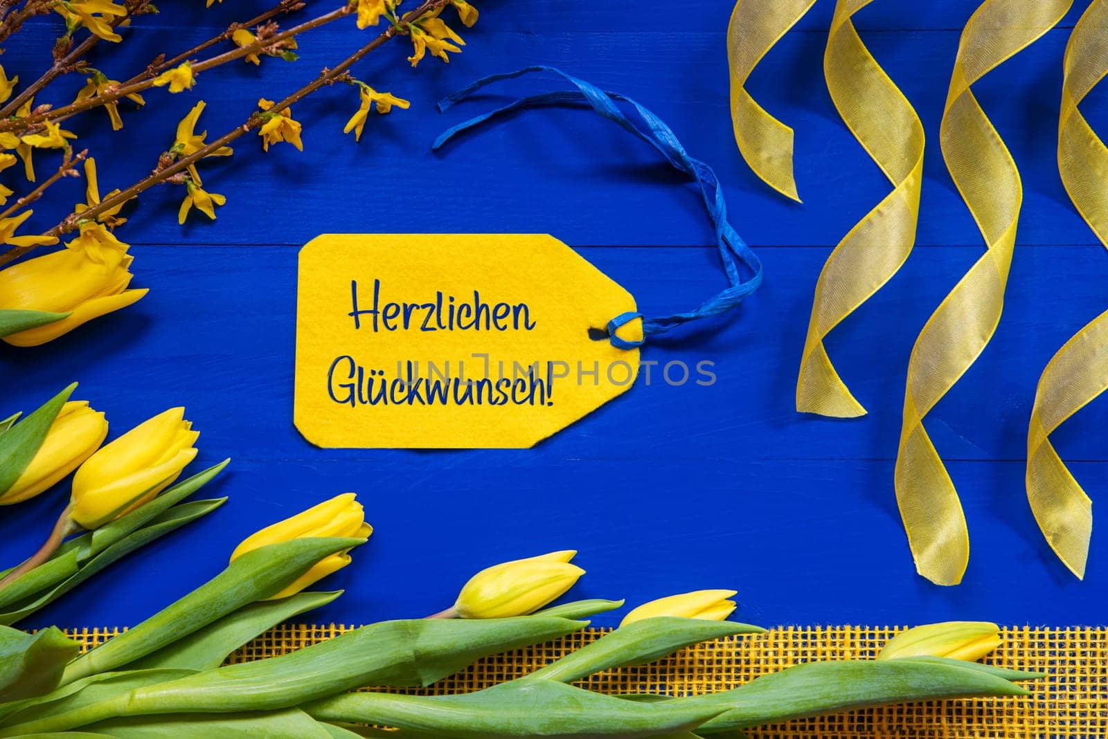 Label With German Text Herzlichen Glueckwunsch Means Congratulations. Yellow Spring Flowers Like Tulip And Branches. Festive Decoration With Ribbon. Blue Wooden Background