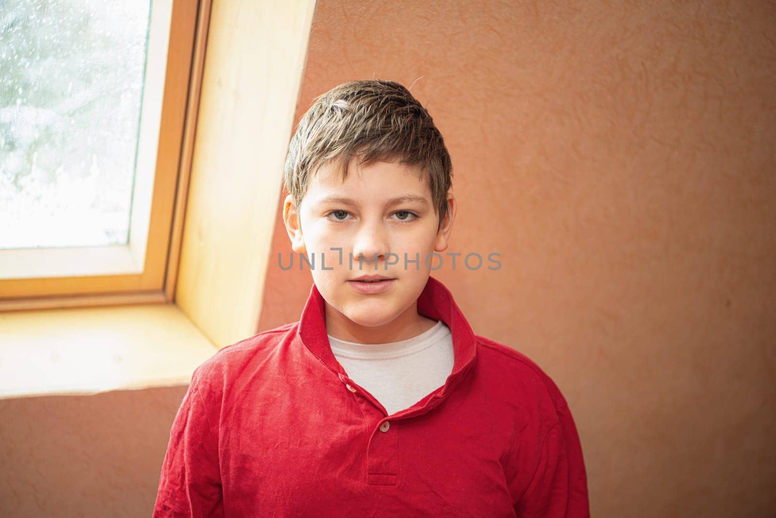 A white boy of school age stands near the window, dressed in a red shirt.