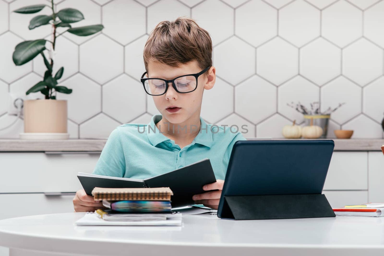 Portrait of young preteen focused boy wearing blue T-shirt, glasses, sitting at desk near notepads, looking at open black book near tablet, having online lesson, reading. E-learning, technology.