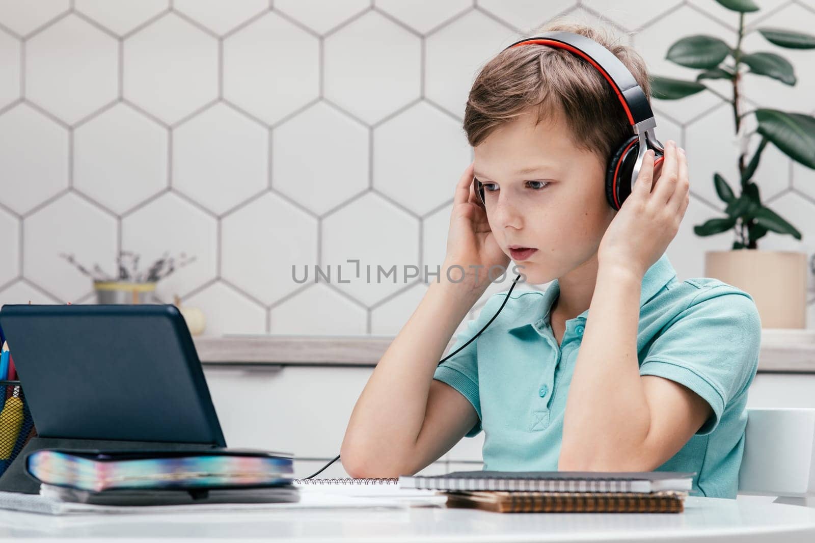 Portrait of young preteen concentrated boy covering headphones with hands, sitting at desk near notebooks at home, looking at screen of tablet, listening to online lesson. E-learning, technology.