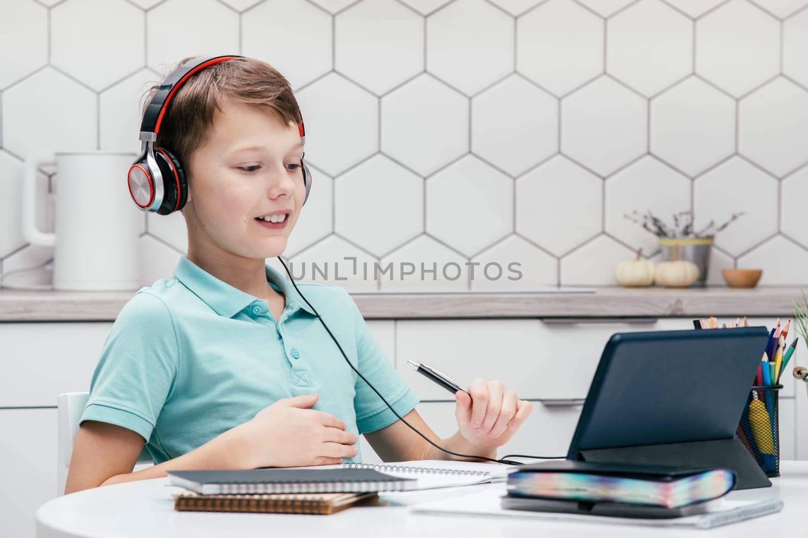 Portrait of young preteen busy boy wearing T-shirt, headphones, sitting at desk near notebooks, looking at screen of tablet, having online lesson, holding pen. Remote education, distance learning.