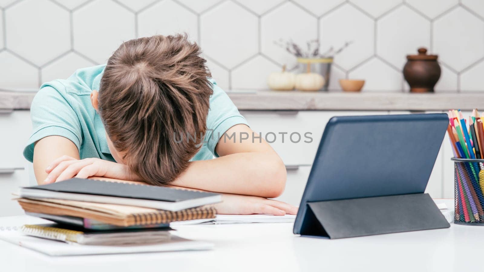Portrait of young preteen tired boy pupil wearing blue T-shirt lying sleeping on folded hands on desk near notebooks, glasses, color pencils, tablet at home. Distant learning, education, technology.