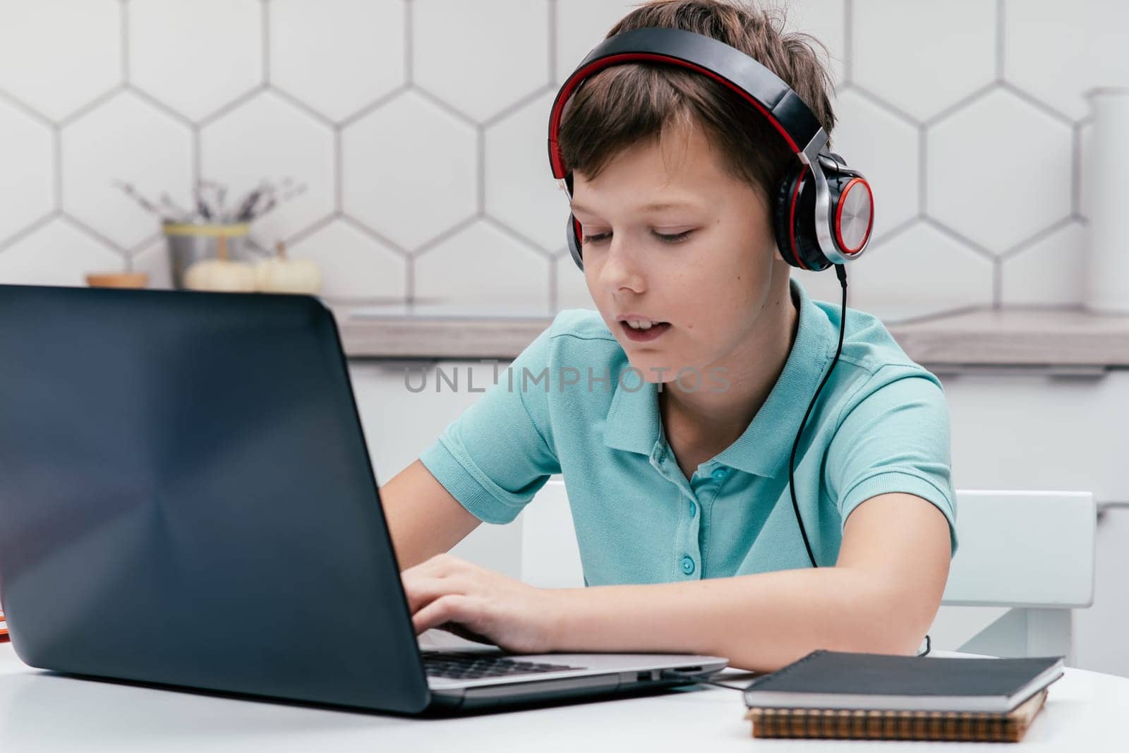 Portrait of young preteen pleased boy wearing T-shirt, headphones, sitting at desk near notebooks, using laptop, having online lesson, messaging. Remote education, distance learning, social media.