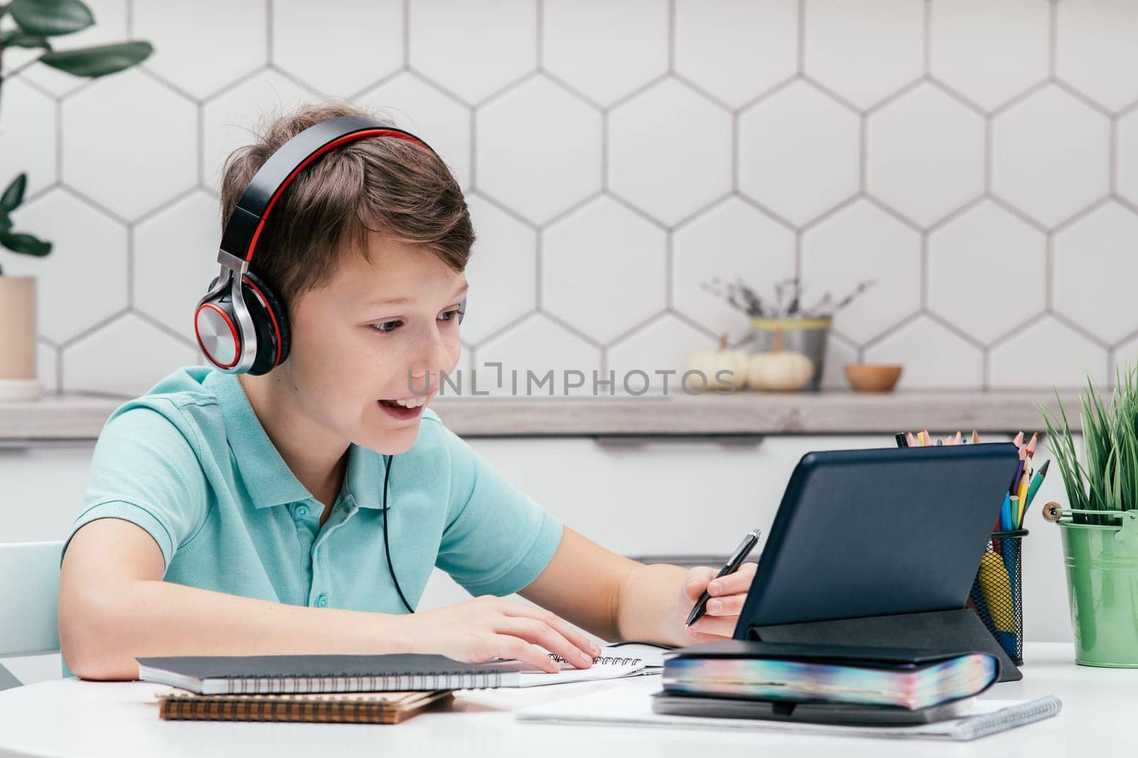 Portrait of young preteen surprised boy wearing headphones, sitting at desk near notebooks, looking at screen of tablet, having online lesson, listening, making notes, holding pen. Distant learning.